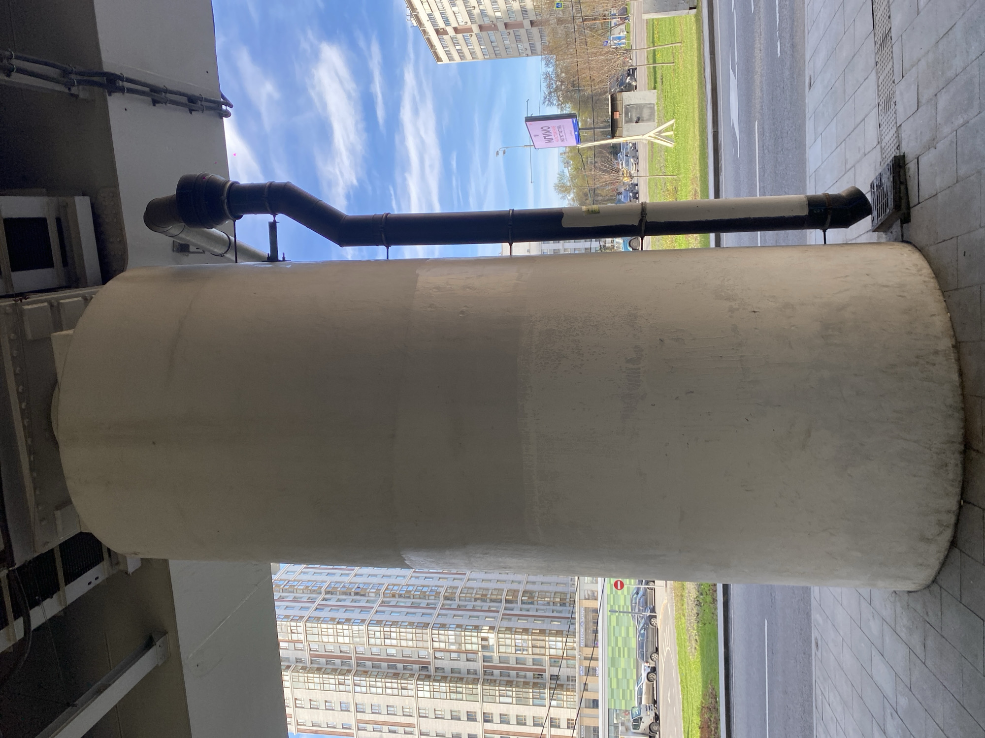 The image captures an urban scene from beneath an elevated structure, likely a bridge or overpass. A prominent, large cylindrical concrete support column dominates the right foreground, extending vertically through the frame. Along its upper section, a series of dark pipes are attached, running horizontally and then angling upwards to connect with the overhead structure. One of these pipes extends towards the right, terminating near a metal grate embedded in the light grey paved ground.
To the left of the column, a multi-lane asphalt road with a distinct white lane marking stretches across the mid-ground, bordered by a strip of vibrant green grass. Several parked cars are visible along the roadside. In the background, tall, multi-story residential or commercial buildings with numerous windows rise against a bright blue sky dotted with scattered white, wispy clouds.
A large digital billboard is visible above the road, displaying text in Cyrillic. The words "МГИМО" (MGIMO), identifying a prominent university in Moscow, and "Искусство" (Iskusstvo), meaning "Art," are clearly legible, confirming the Moscow, Russia, location context. A red circular sign, typically indicating "No Entry" for vehicles, is also present near the parked cars. The scene is illuminated by natural daylight, suggesting a clear day. No people or specific activities are visible, presenting a static snapshot of city infrastructure.