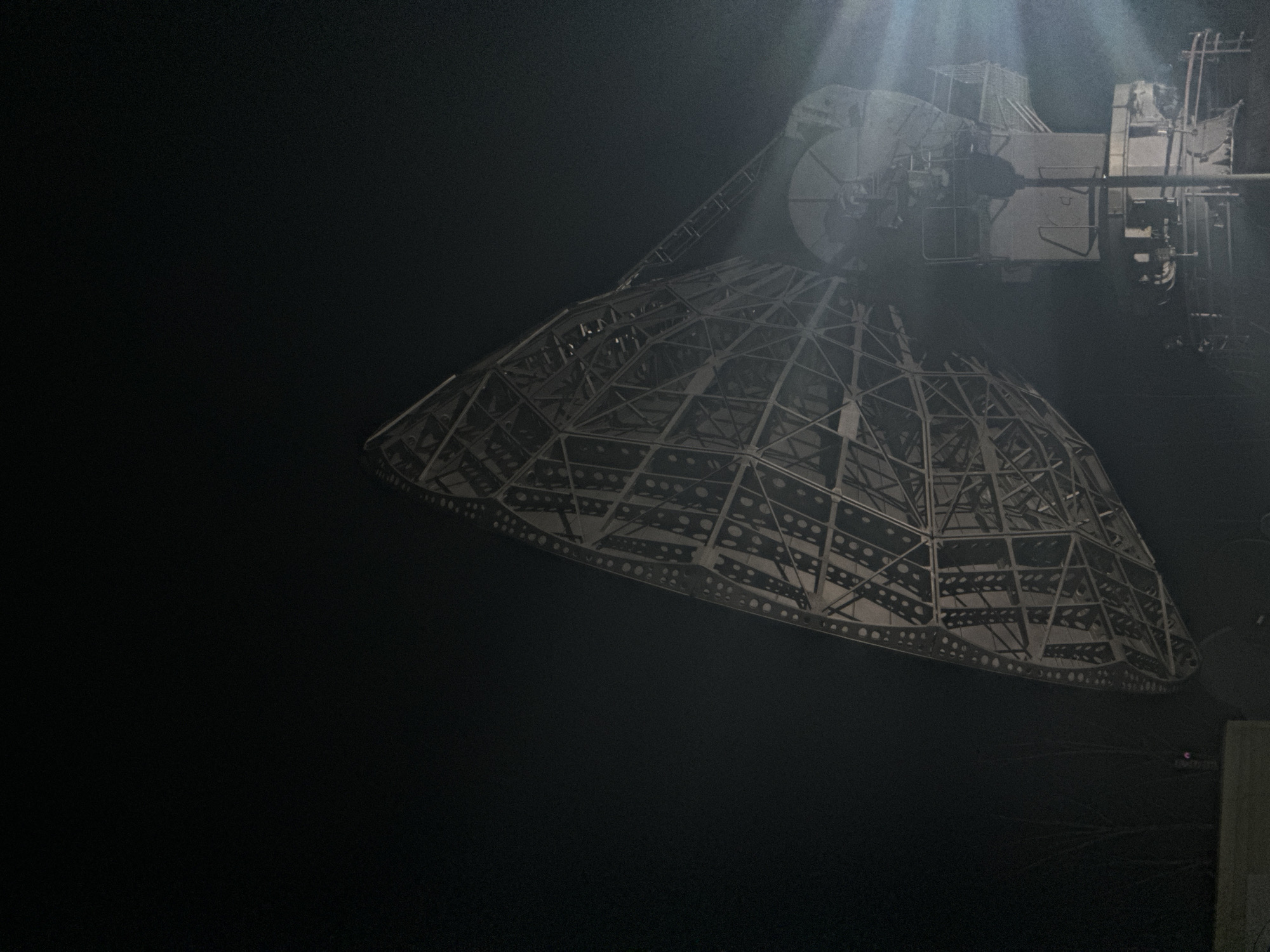 The image shows a large radio telescope against a dark background. The telescope's dish is made of a lattice structure, and its supporting machinery is visible above it. Light shines down on the telescope from above. The location of the telescope is in the city of Krasnoyarsk, Russia.