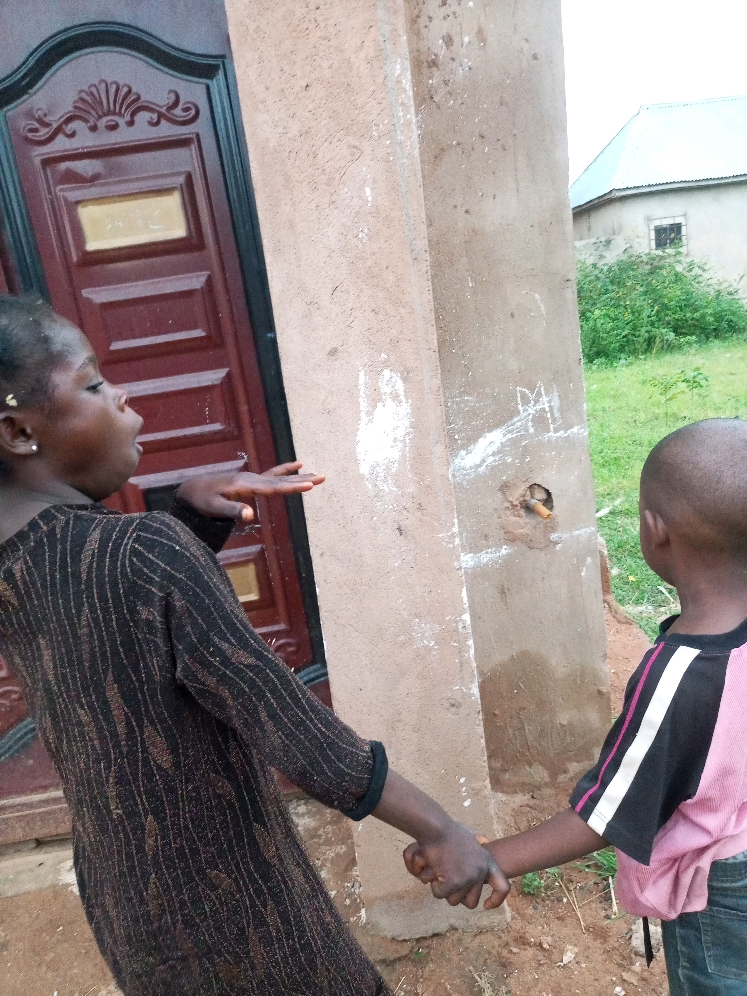 Two young children are standing in an outdoor environment. The child on the left, facing away from the camera and to the right, has dark skin and is wearing a dark, patterned long-sleeved dress. Their left hand is extended outwards towards a concrete pillar, while their right hand is clasped with the child on the right. The child on the right is facing away from the camera and to the left, and has a shaved head. They are wearing a pink shirt with black and white stripes on the sleeves and dark pants. The children are holding hands, suggesting an interaction or relationship between them.
The environment appears to be semi-urban or rural, with a reddish-brown dirt ground, some dry grass and vegetation visible in the background. To the left of the children is a dark red door with decorative elements and a rectangular insert. To the right is a rough, textured concrete pillar with some white markings and a small, circular opening with a piece of what appears to be a cigarette butt protruding from it. In the background, a light-colored building with a light blue or grey roof is visible, along with some green bushes. The overall lighting suggests it is daytime, possibly overcast. The image has a slightly grainy quality.