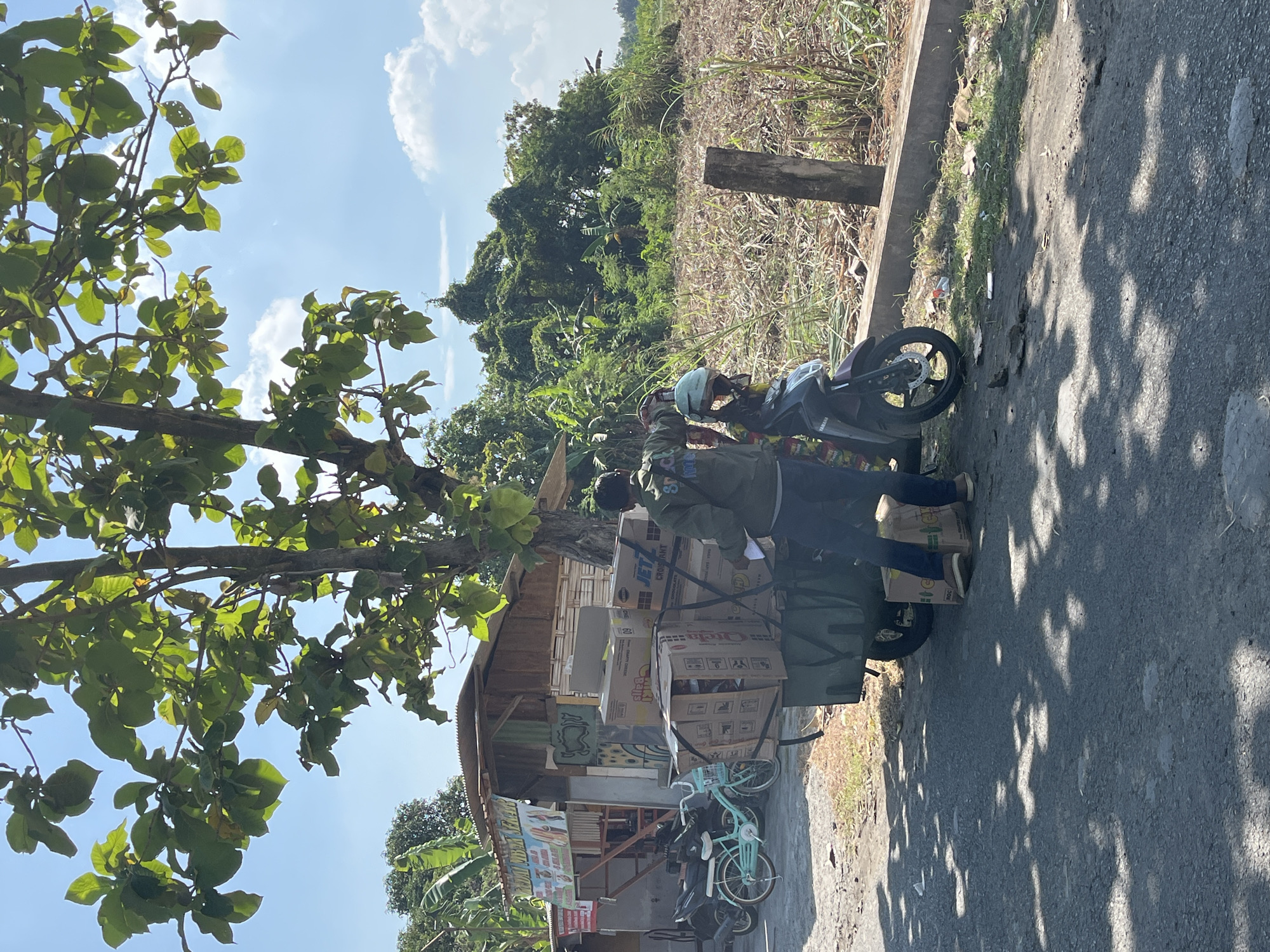 In Pamotan, Indonesia, a man in a green jacket and dark pants stands next to a motorcycle with a trailer filled with boxes. The trailer is attached to the motorcycle's rear wheel. The motorcycle also carries a helmet. The man appears to be in the process of handling the boxes. In the background, there is a small wooden building with a corrugated metal roof, possibly a shop, and a tree with large green leaves. Beyond the building and the road, there is a field with dry vegetation and a dense area of green trees. The sky is blue with scattered white clouds. A light blue bicycle is parked to the left of the trailer. The scene is lit by sunlight, casting shadows on the asphalt road.