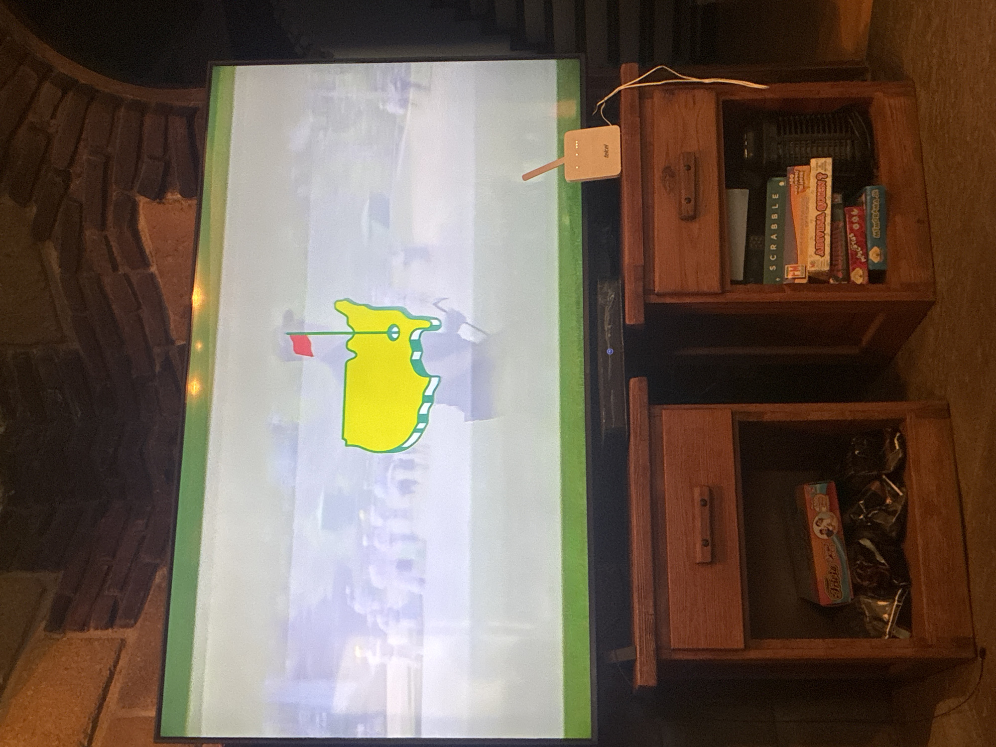 The image shows a television displaying a graphic of the United States in yellow and green. Next to the television are two wooden cabinets, one stacked on top of the other. The top cabinet contains board games, including Scrabble and Trivial Pursuit. The bottom cabinet contains a Trivial Pursuit game and some wrapped items. The setting appears to be indoors. The location in the city of Cañada de Madero, Mexico, is unknown.