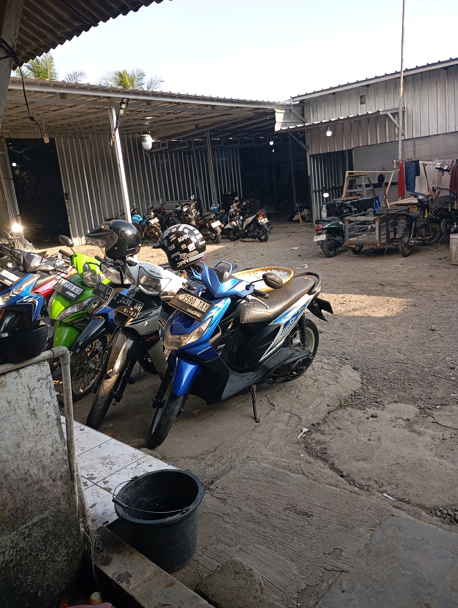The image depicts a street scene in Tasikmalaya, Indonesia, likely an outdoor parking area or workshop. Several motorcycles are parked, with a prominent blue scooter in the foreground. A black bucket sits on tiled ground to the left. The environment is characterized by a corrugated metal roof structure covering a portion of the area, with a gravel and concrete ground surface. The lighting suggests it is daytime, with sunlight casting shadows. In the background, more motorcycles are visible, along with some structures that appear to be part of a commercial establishment. There are no people clearly visible in the frame, though the presence of parked vehicles suggests recent activity. The scene is utilitarian and informal, typical of many urban settings in Southeast Asia.