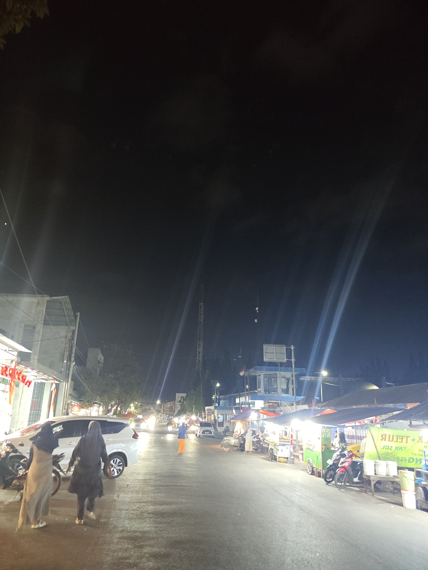 The image depicts a street scene at night in Situbondo, Indonesia, likely a market or food stall area. The street is wet, suggesting recent rain. On the left, a white car is parked, with two women in hijabs standing nearby. One woman, on the left, is wearing a beige dress, and the other, on the right, is wearing a dark abaya. A motorcycle is parked behind the car. Further down the street, there are several food stalls illuminated by bright lights. One stall has a green sign with Indonesian text. Several motorcycles are parked in front of the stalls, and a person in an orange jacket is walking away from the stalls. In the background, there are buildings and a tall radio tower. The sky is dark, with streaks of light emanating from what appear to be spotlights. The overall atmosphere is that of a bustling night market.