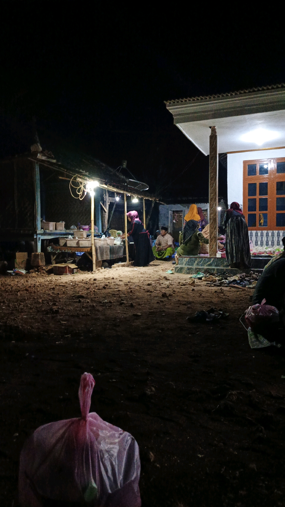 At night in Sokaramme Timur, Indonesia, a scene unfolds outdoors near a dwelling. In the foreground, a pink plastic trash bag is visible. Further back, a simple stall or shop is illuminated by overhead lights, displaying various items in baskets on a table. Several individuals are gathered in and around this area. One person, wearing a dark abaya and headscarf, stands near the stall, possibly interacting with someone or arranging items. Another individual, a man in a light-colored shirt and dark trousers, sits on the ground. A woman in a bright yellow headscarf and green garment is seated on steps. Adjacent to the stall, a house with a white facade and a wooden-framed window is partially visible. More people are gathered near the house, with one person in a dark, patterned abaya standing. The ground is unpaved and appears to be dirt or sand. The overall impression is of a communal gathering or market scene taking place after dark.