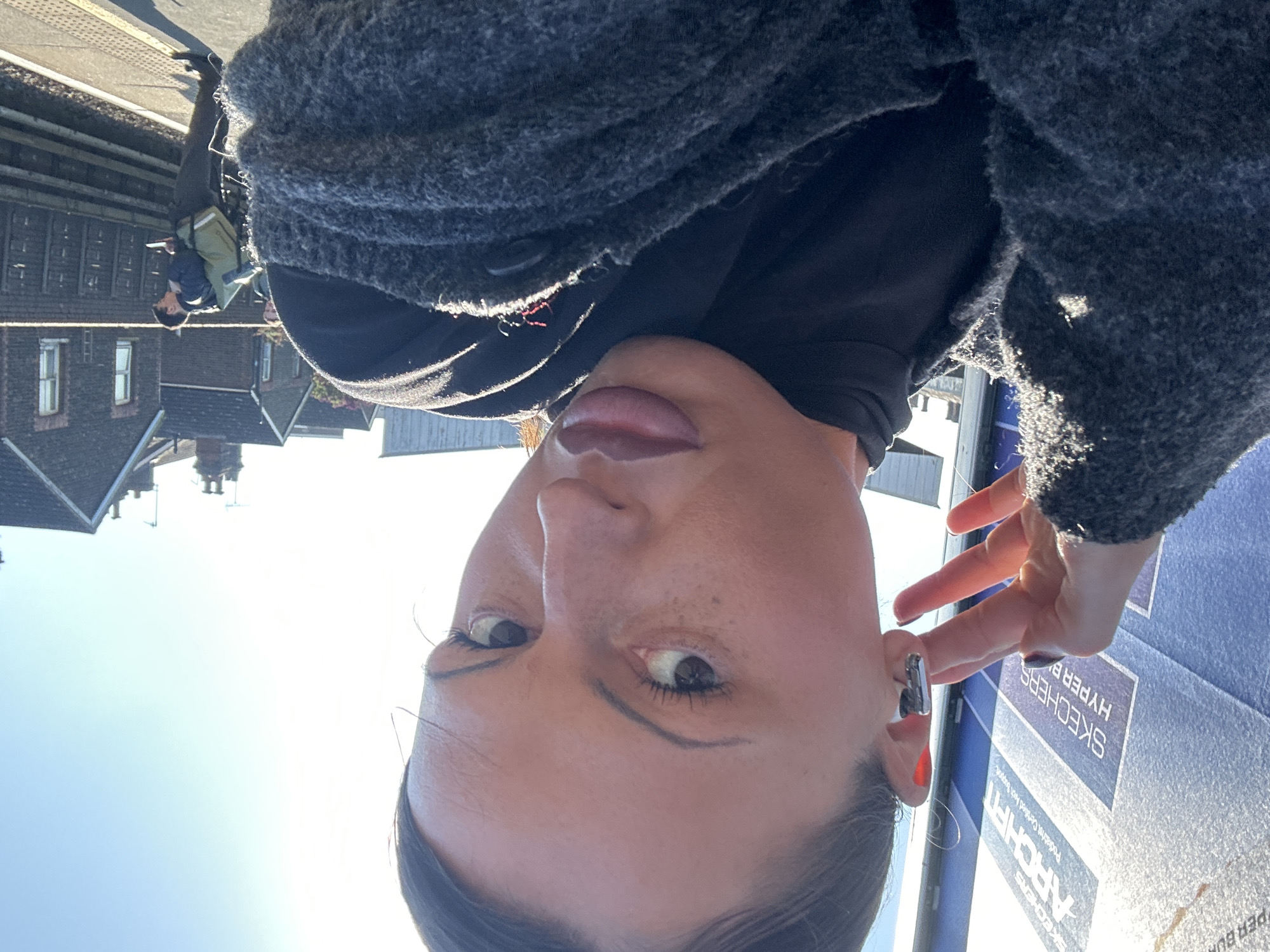 A bright outdoor selfie captures a woman from a slightly low angle, positioned in the foreground. She has light brown skin with visible freckles, dark hair, and is wearing eyeliner and reddish-pink lip color. Her eyes are light-colored, and she looks directly towards the camera with a neutral expression. She wears a textured dark grey cardigan or jacket over a black top. A metallic earring or earbud is visible in her right ear, and her fingers gently touch her earlobe.
The background reveals a sunny day with a clear blue sky. On the right, a large blue sign is partially visible, displaying white text that reads "SKECHERS," "HYPER B," "ARCH FIT," and "Product Certified By Experts." To the left, a train platform or paved area stretches into the distance, where a person, possibly a man or boy, is walking away from the camera, carrying a khaki or light green backpack. Further back, several buildings with dark roofs and windows are visible, suggesting an urban or suburban setting. The bright sunlight casts strong shadows and highlights across the scene, indicating a mid-morning or afternoon time of day.