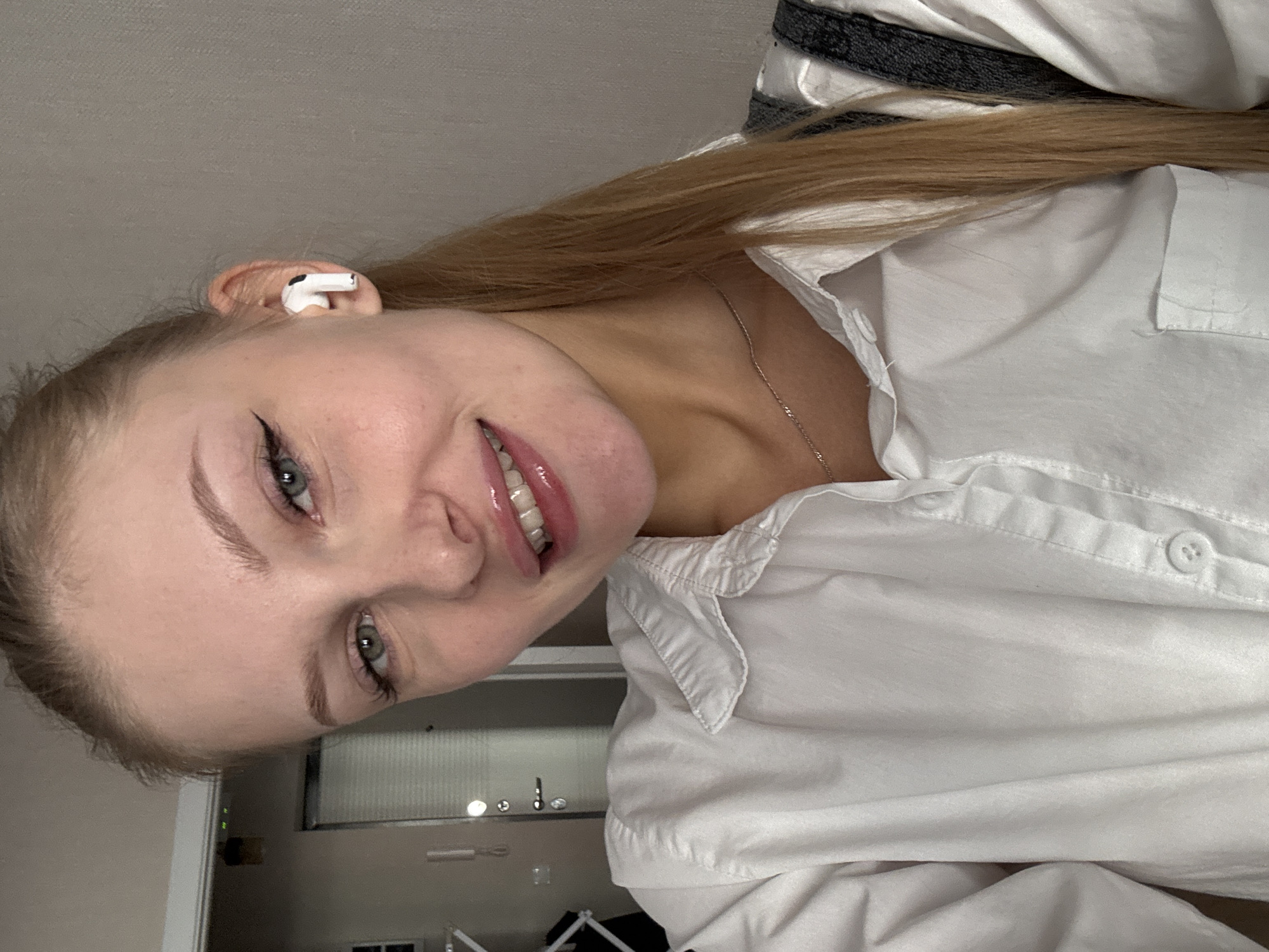 A young Caucasian woman with blonde hair pulled back in a ponytail and green eyes is looking directly at the camera and smiling. She is wearing a white button-down shirt and white wireless earbuds. The background appears to be an indoor setting with a wall and a doorway visible. The image is a close-up shot, focusing on the woman's face and upper body. There is no indication of the city of Perm, Russia.