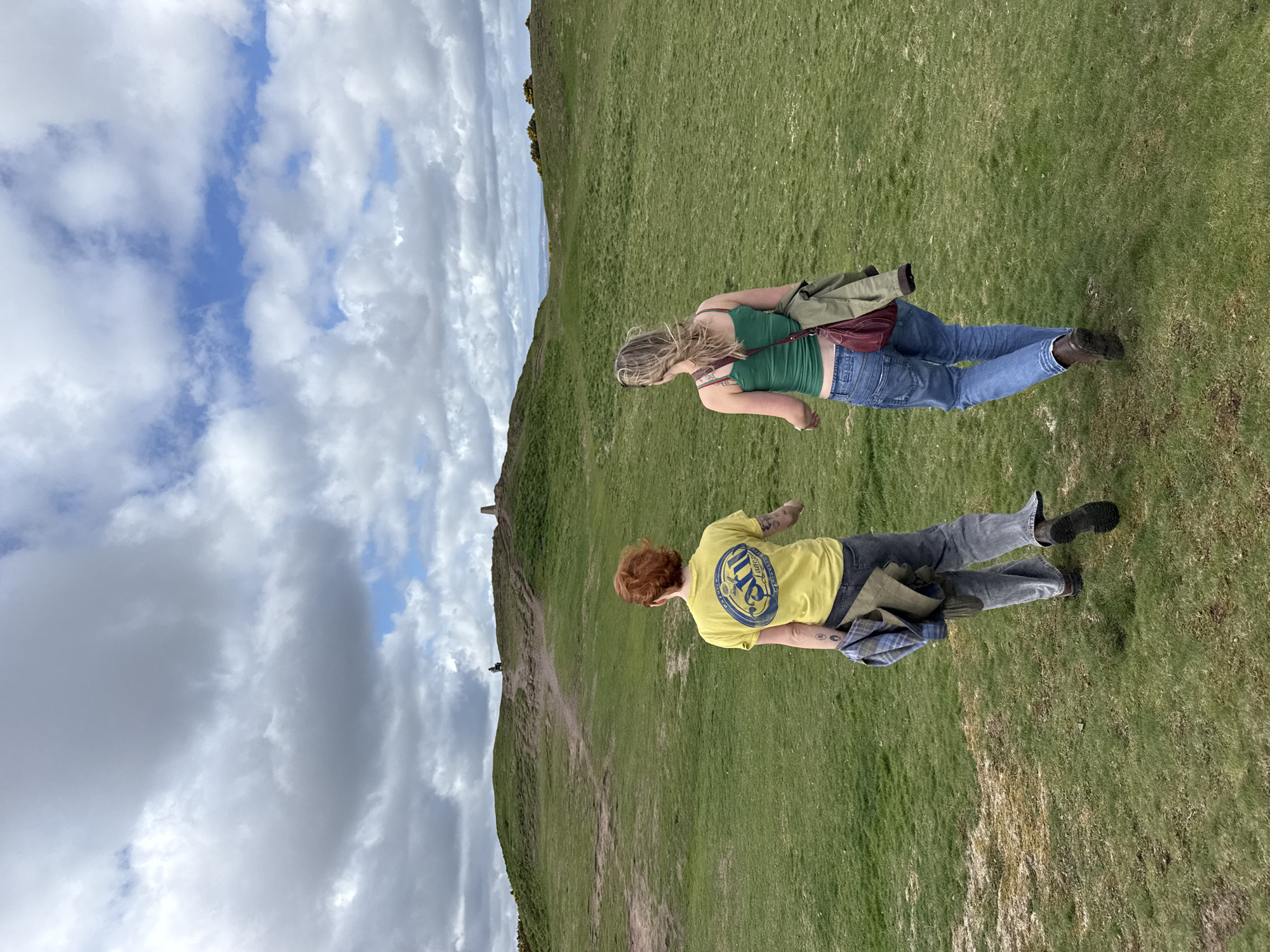 Two individuals are walking on a vast, green grassy hillside under a partly cloudy sky during the daytime. The image is oriented vertically, showing the people standing upright relative to the ground.
The person in the foreground has red hair, is wearing a yellow short-sleeved t-shirt, blue jeans, and a casual bag slung across their shoulder. The yellow t-shirt features a prominent blue circular logo on the back, which appears to be for "MILLER" beer, possibly "MILLER HIGH LIFE."
Further up the slope and slightly behind, a second person with blonde hair is seen wearing a green tank top, blue jeans, and a maroon fanny pack or cross-body bag around their waist, with a khaki jacket draped over it.
The terrain consists of a gently sloping green hill, with a visible dirt path winding up towards the crest. In the distance, a small, dark structure, possibly a monument or cairn, is perched on the hilltop. The sky above is a mix of white clouds and patches of blue. The overall impression is of a casual walk or hike in a natural, open landscape. The weather appears bright but with significant cloud cover.