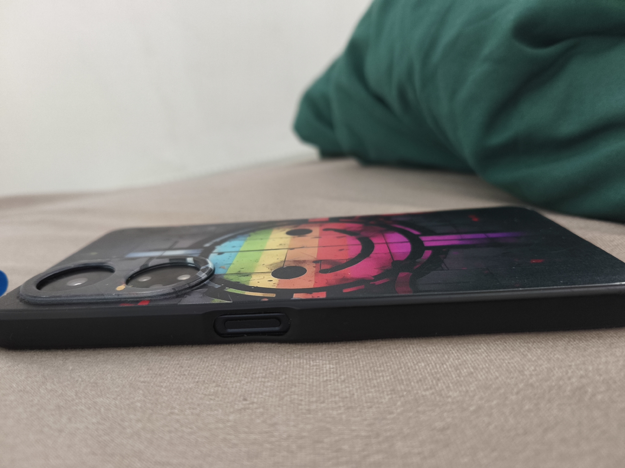 A smartphone with a colorful smiley face design is lying on a textured surface. The phone is black with a case that features a rainbow-colored smiley face with headphones. The camera lenses are visible on the back of the phone. The phone is angled, showing its side profile. In the background, a crumpled green fabric is visible, suggesting it might be a blanket or clothing. The overall scene is a close-up shot of a mobile device.