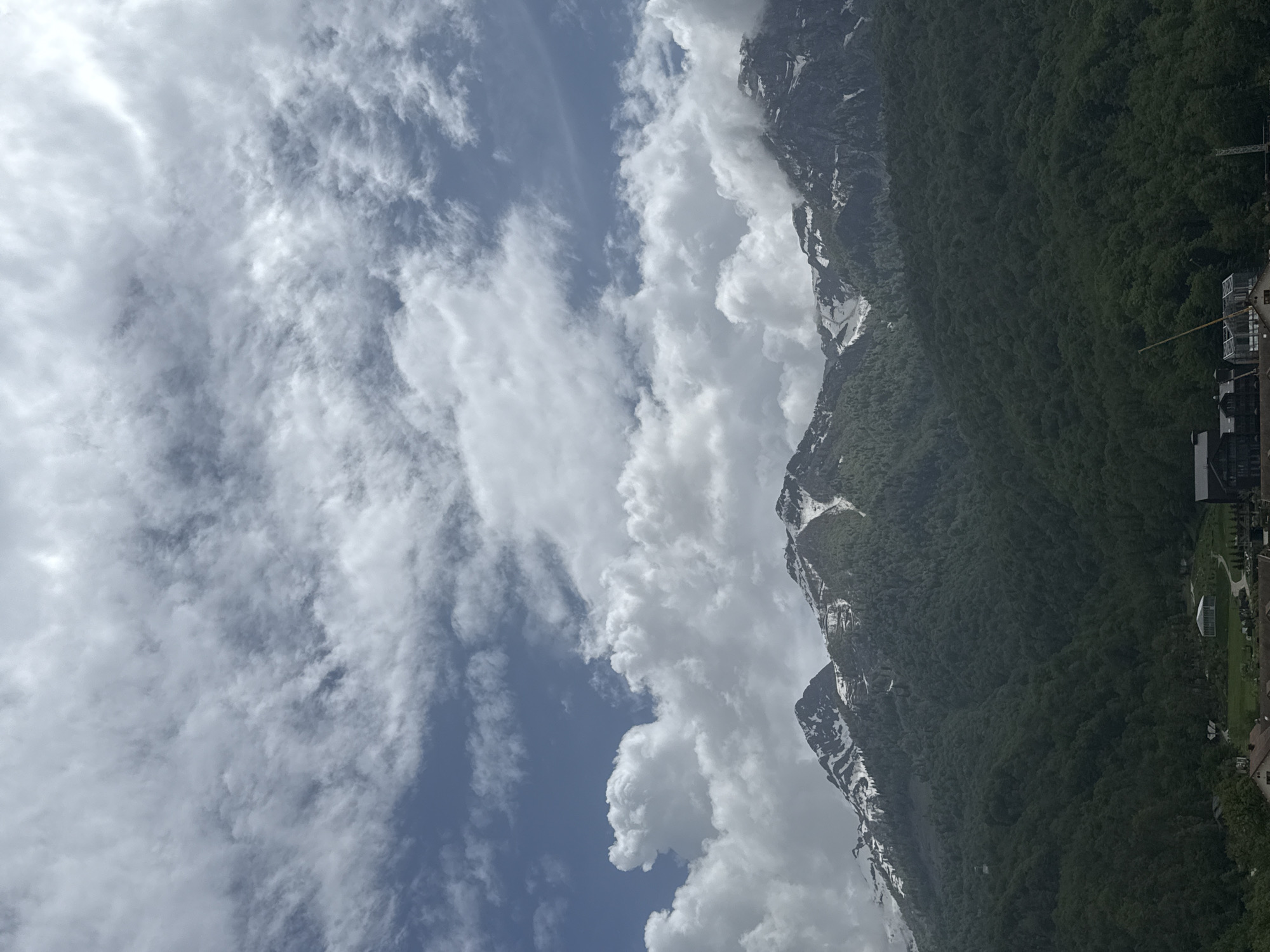 The image shows a landscape in Krasnaya Polyana, Russia, featuring a mountain range with snow patches, densely forested slopes, and a cloudy sky. A structure resembling a gondola lift is visible on the right side of the image, along with some buildings below. The weather appears to be partly cloudy, with some blue sky visible amidst the clouds.