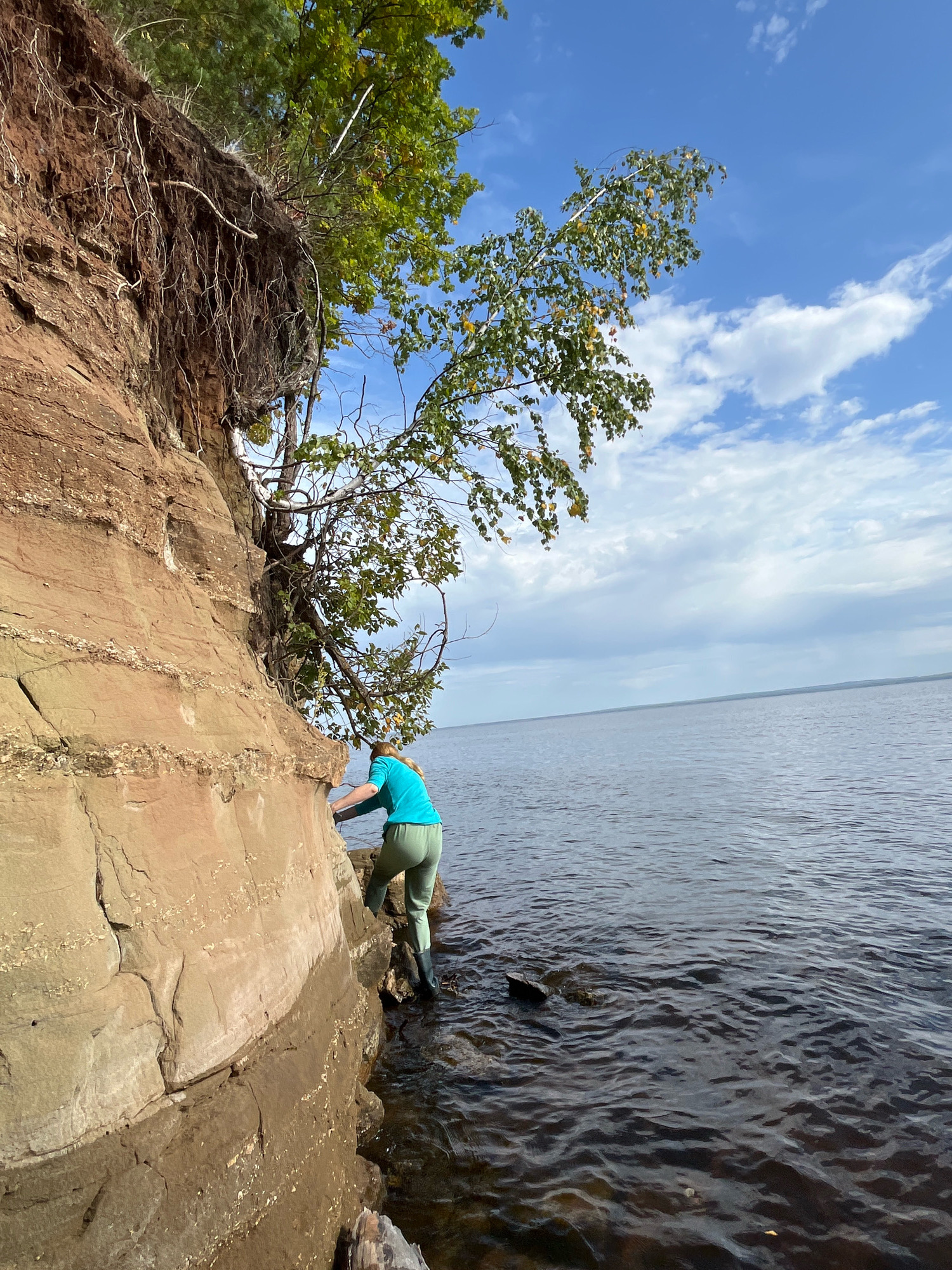 A person wearing a turquoise shirt and green pants climbs on a rocky cliff face next to a body of water. The cliff is made of layered, sandy-colored rock with exposed roots from trees growing from the top. The water is dark and appears to be a lake or river, with ripples on the surface. The sky is blue with scattered white clouds. The scene appears to be a natural landscape, possibly along the Kama River in Chaikovsky, Russia, given the description provided of the location. The person is actively engaged in climbing the rock face.