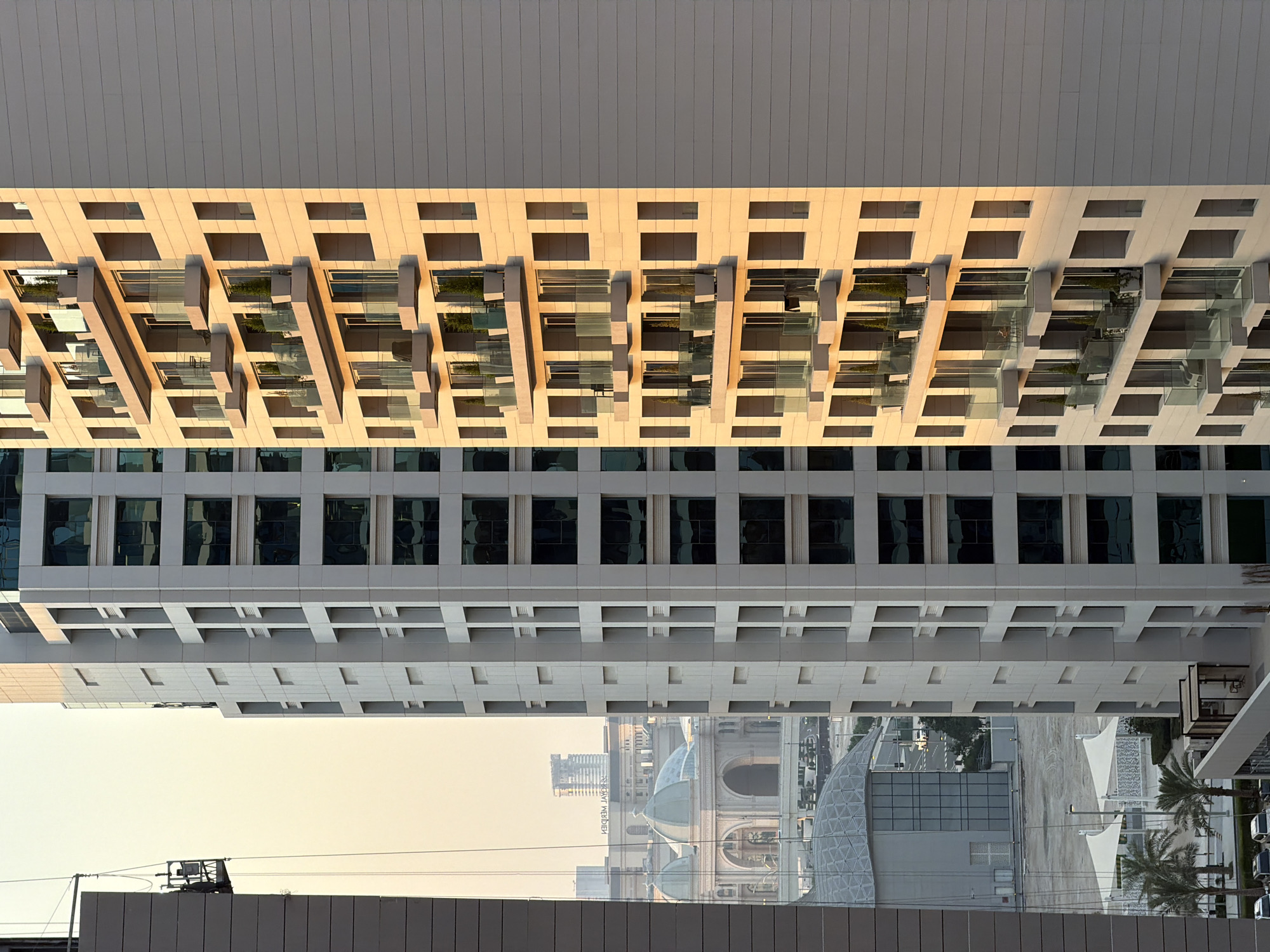 The image shows a low-angle shot of modern architecture, likely an office building or apartment complex, bathed in the warm light of either sunrise or sunset. The building's facade features a repetitive pattern of balconies with glass railings and dark window frames, interspersed with lighter colored geometric protrusions. The upper section of the building is a flat, light-colored surface with horizontal lines. Below this, another section of the building displays rows of dark, rectangular windows. In the distance, the reflection in the windows suggests a city skyline with domes and other structures. The sky is a pale yellow, indicating a specific time of day. The image is presented upside down. There is no discernible text.