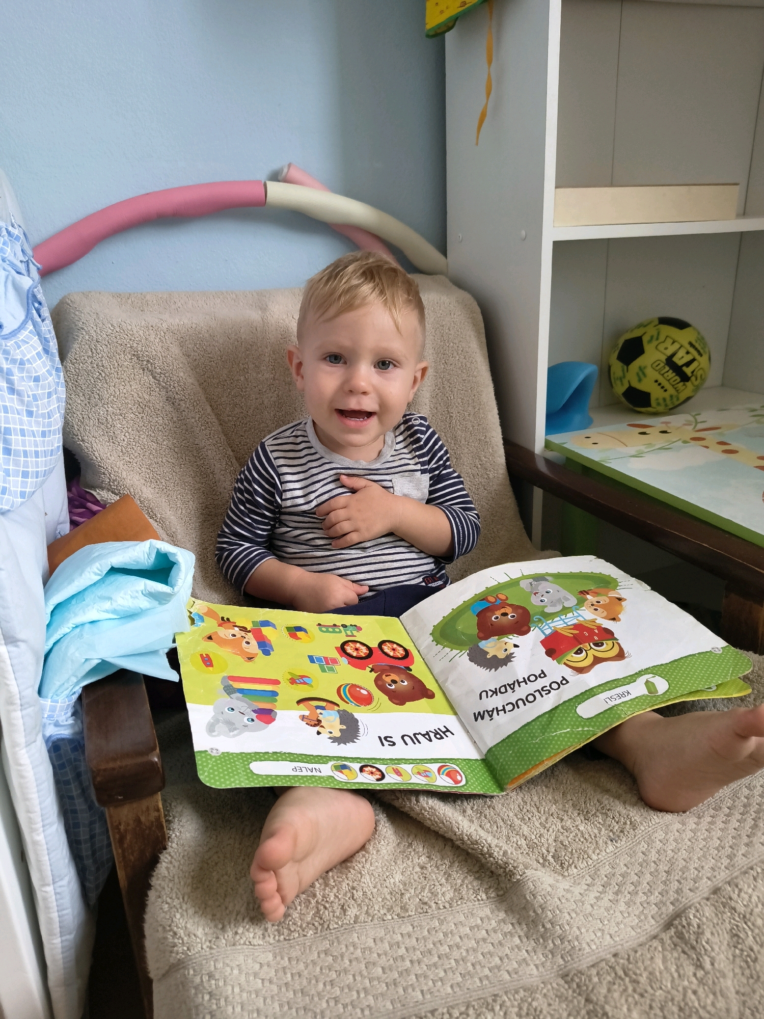 A blond-haired Caucasian toddler sits in a chair covered with a tan blanket, holding an open children's book. The book features colorful illustrations of animals and text. The toddler is wearing a striped long-sleeved shirt and dark pants. Their left hand is on their chest, and they are looking at the camera with a smiling expression. In the background, there is a light blue wall with a pink and cream-colored hula hoop leaning against it. To the right, a white shelving unit is visible, holding a yellow and black soccer ball and a mat with a giraffe illustration. The scene appears to be indoors, possibly in a home environment in Olomouc, Czechia.
