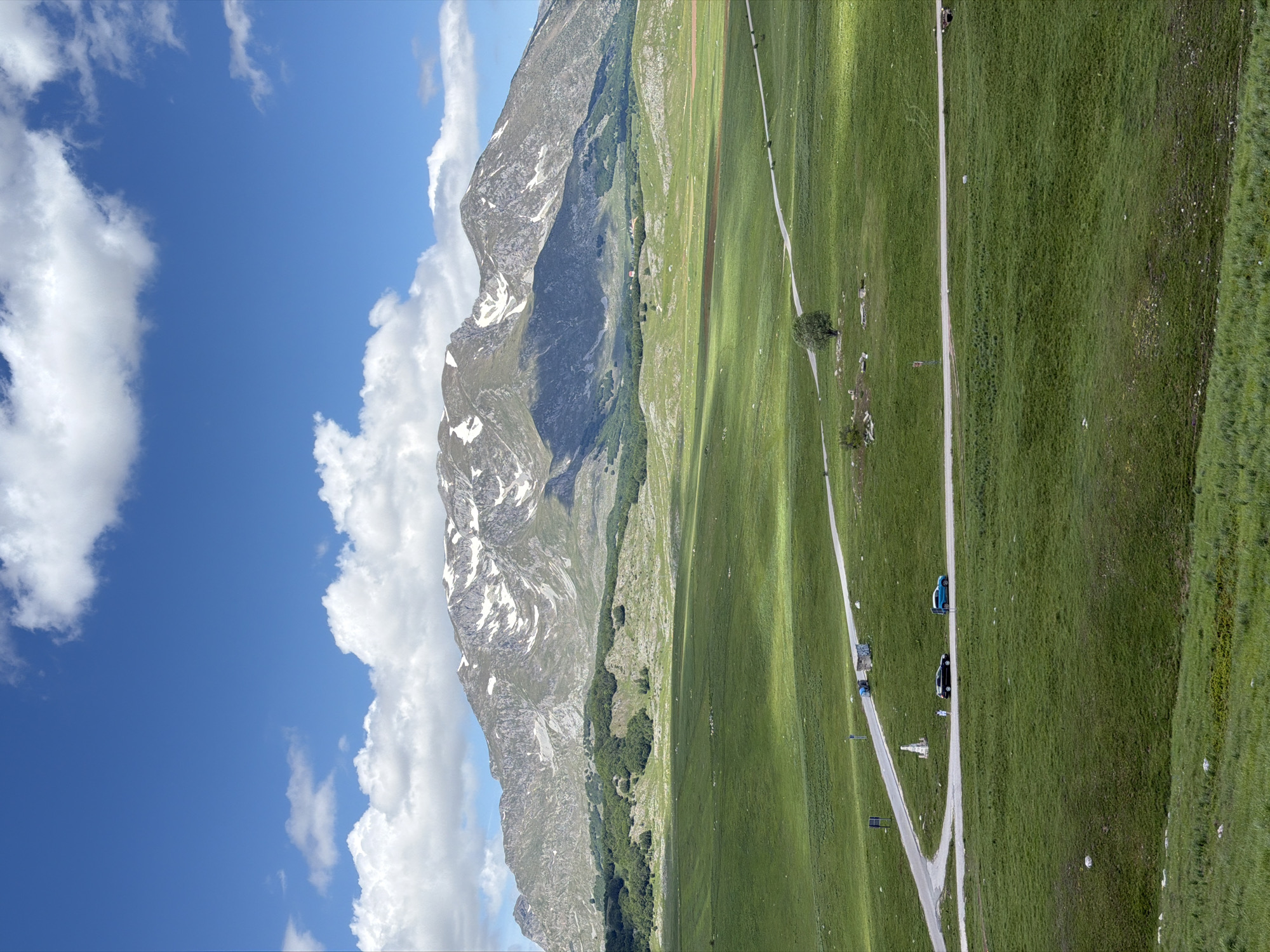 This image captures a vast, open landscape in Novakovići, Montenegro, under a bright blue sky partially covered by white cumulus clouds. In the background, prominent rocky mountains with patches of residual snow dominate, exhibiting grey and brown tones with some green vegetation on their lower slopes. Below the main mountain range, a series of undulating hills covered in dense green foliage, consistent with forests or dense shrubbery, transitions into expansive, bright green grasslands that fill the midground and foreground.
A paved asphalt road winds through these verdant fields in the mid-foreground, forming a distinct 'Y' intersection or sharp bend. At this junction, a white stone monument or pillar stands by the roadside. Two passenger vehicles are present on the road: a dark-colored car, possibly black, parked or moving slowly, and a light blue car further along the road. A solitary person, dressed in white, is visible standing near the dark-colored car. Further down the road, a small, light-colored building or structure is situated, accompanied by two blue waste or recycling bins. Scattered road signs are also visible alongside the road. A single, well-formed deciduous tree with a full green canopy stands isolated in the field to the right of the road junction. Sunlight casts clear shadows, indicating a bright day, likely midday or early afternoon.