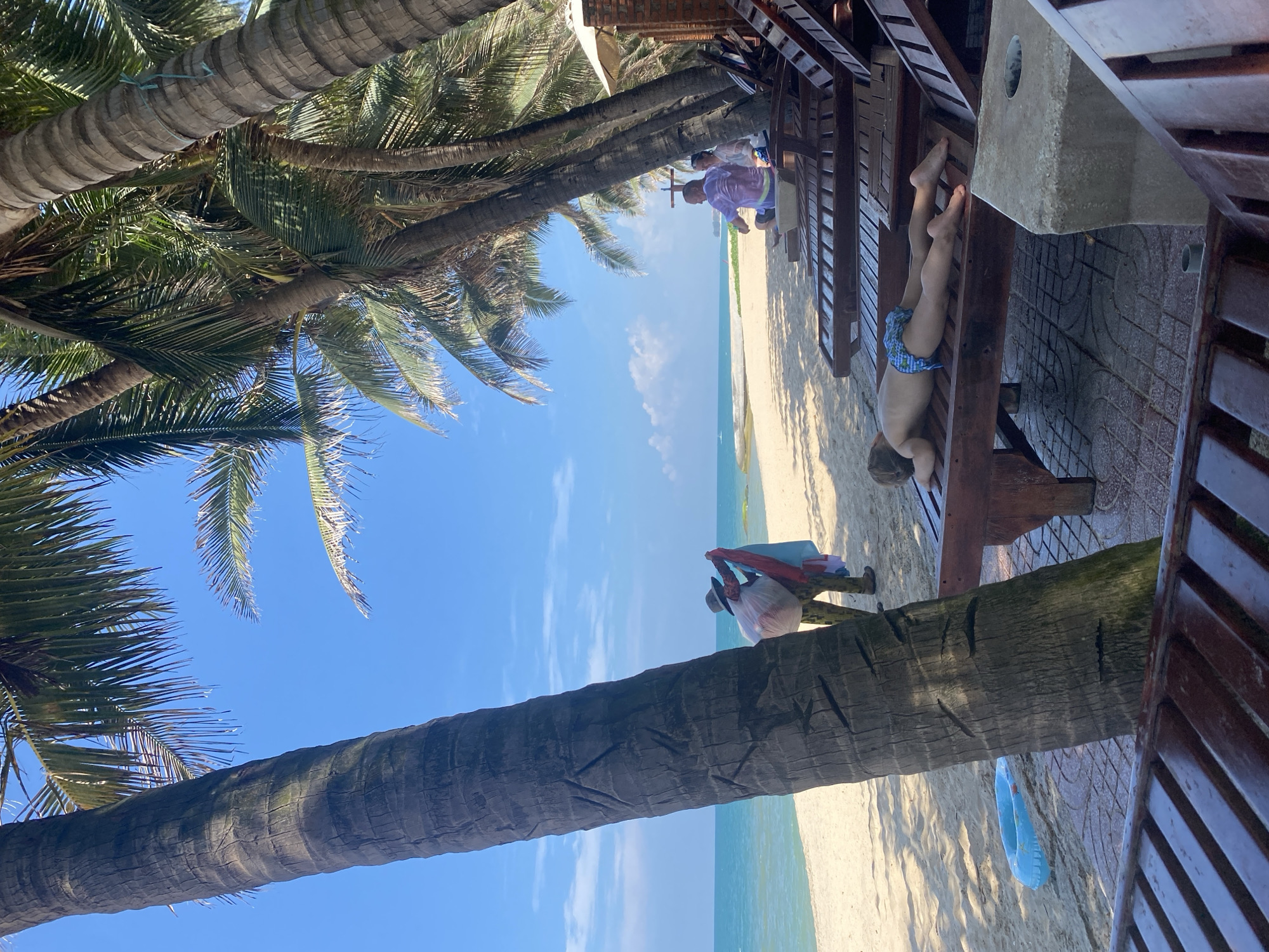 The image depicts a beach scene with several palm trees lining the shore. In the foreground, wooden lounge chairs are set up on a tiled area. One lounge chair in the mid-ground has a young child lying face down on it, wearing blue patterned swim trunks. Further back, a person wearing a hat and carrying laundry is standing near a palm tree on the sandy beach. The ocean is visible in the background under a clear blue sky with scattered clouds. Two adults are seated on lounge chairs further back, under the shade of the palm trees, facing the ocean. The overall atmosphere suggests a relaxing day at the beach. The location is identified as Phu Thuy Ward, Vietnam.