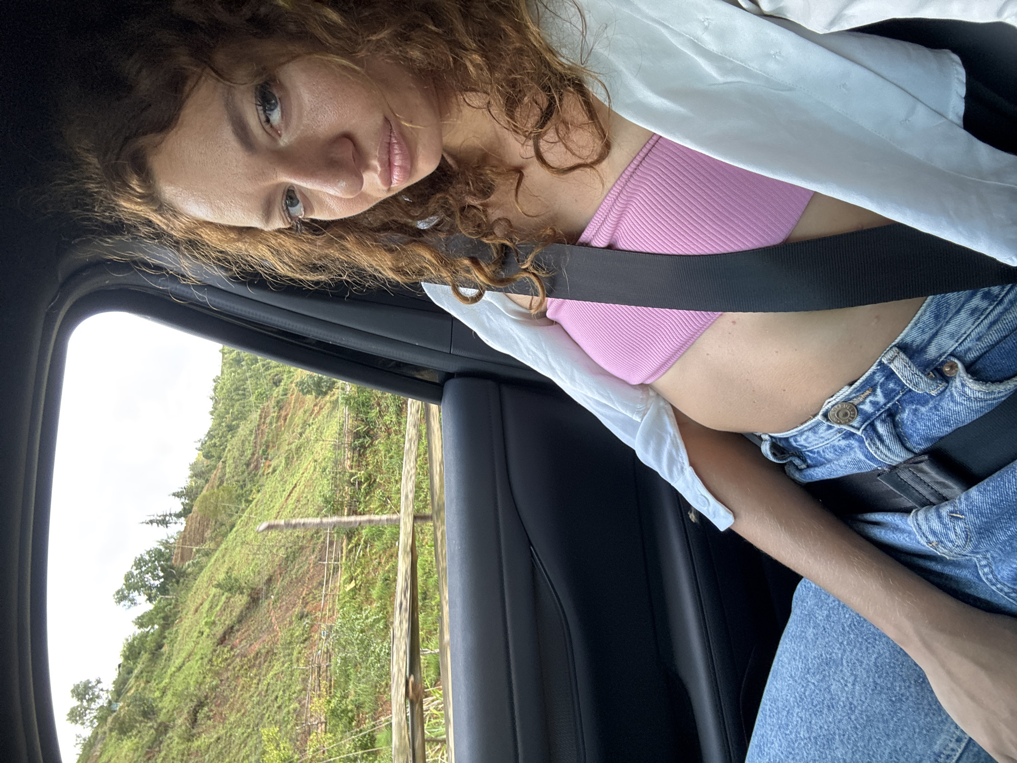 A young woman is sitting in a car, wearing a pink top, jeans, and a white shirt with the seatbelt fastened. She has long, curly hair and is looking at the camera. The car is passing a hillside with trees and vegetation. The location is Ngã Ba Kim, Vietnam.