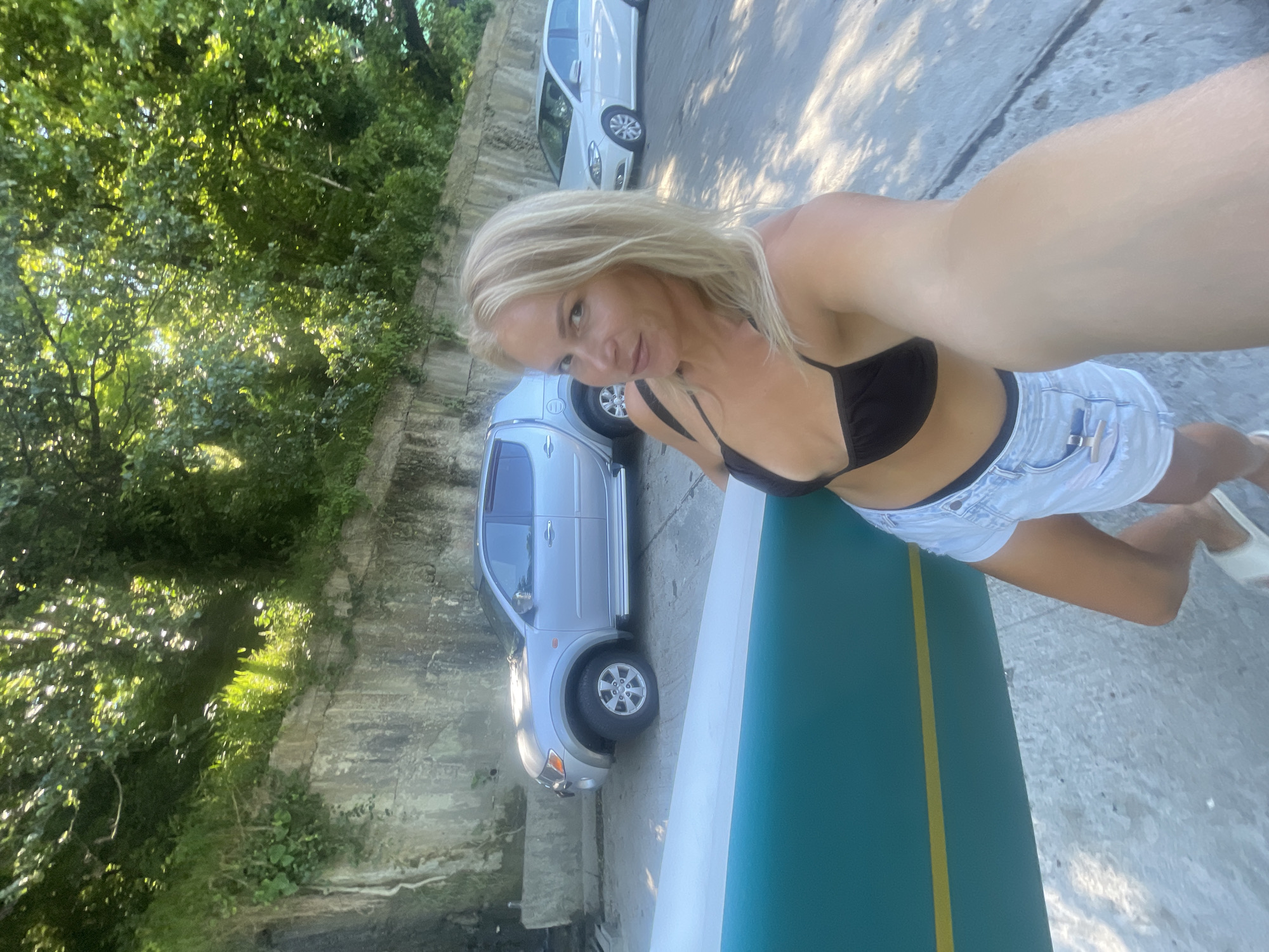 A blonde woman in a black bikini top and denim shorts is taking a selfie while holding a surfboard. She is standing on a paved surface next to a concrete wall with trees and foliage above it. Behind her, a silver pickup truck and a white car are parked. The sun is shining, casting shadows on the ground. The scene appears to be in a park or near a beach in Sochi, Russia, given the vegetation and the presence of a surfboard.