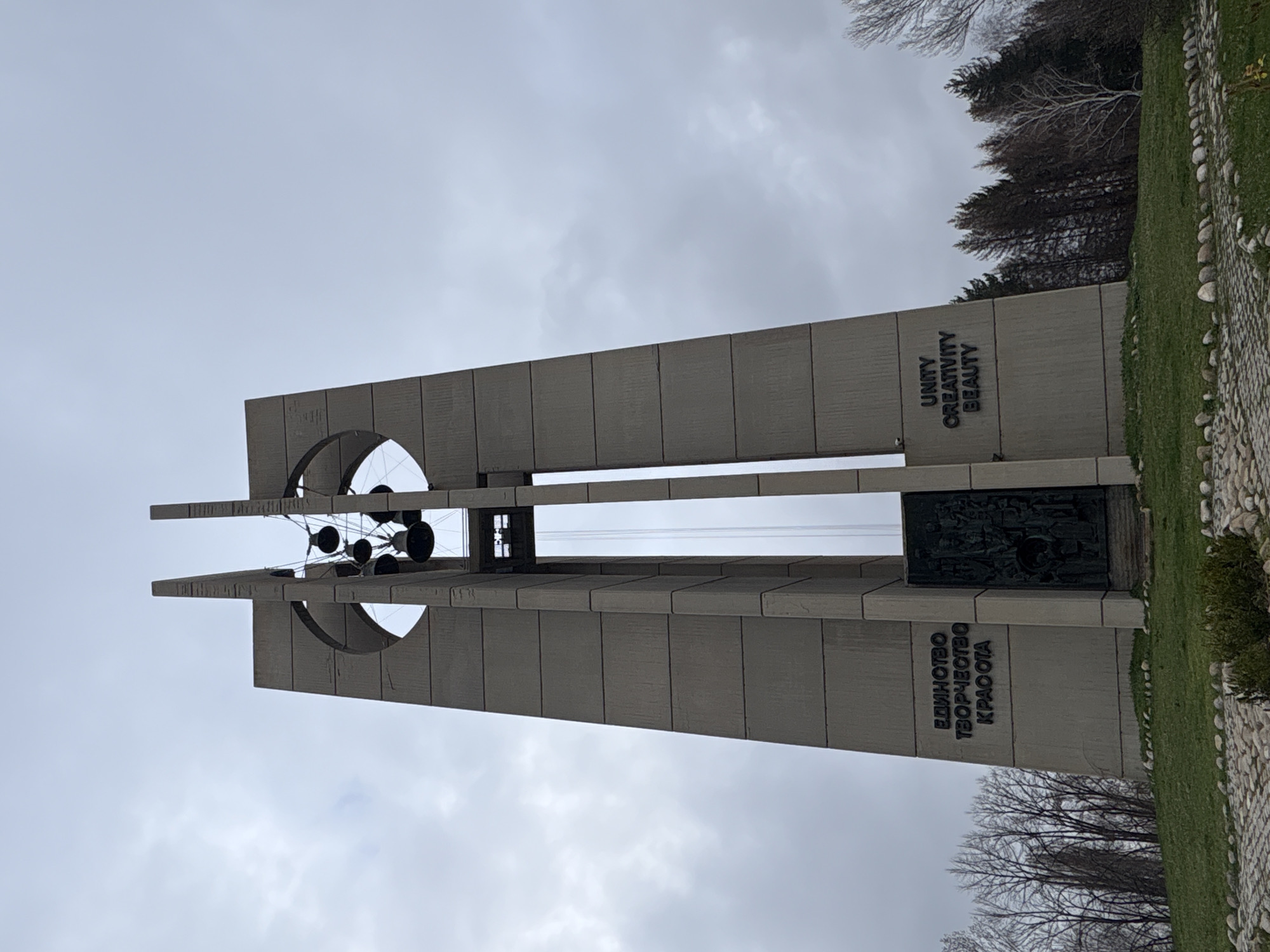 The image shows the Bells Monument in Sofia, Bulgaria, a tall concrete structure with two towers. The towers have circular cutouts and metal bells suspended in the center. One tower has the inscription "UNITY CREATIVITY BEAUTY" in English, while the other has the same inscription in Bulgarian ("ЕДИНСТВО ТВОРЧЕСТВО КРАСОТА"). A dark relief panel is visible on the side of the monument. The surrounding environment consists of grass, trees, and a pebble border. The sky is overcast.