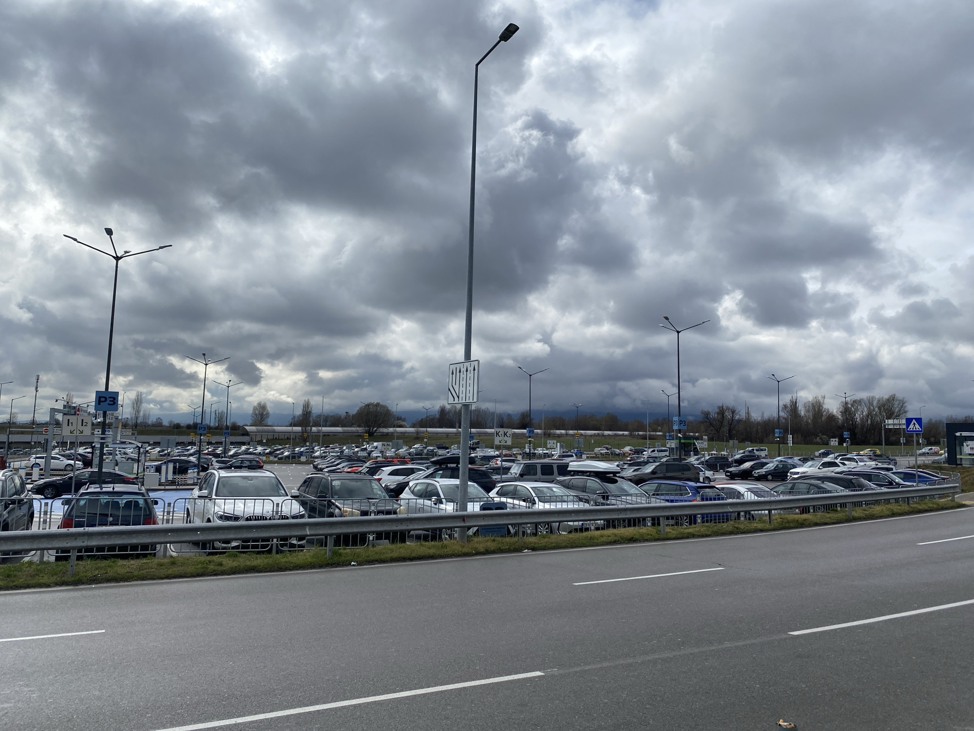 The image depicts a large parking lot filled with cars under a cloudy sky. The location is the Sofia Airport parking lot in Sofia, Bulgaria. The parking lot is separated from the road by a metal barrier and a strip of grass. The road in the foreground is asphalt with white lane markings. Several lampposts are visible throughout the parking lot. The sky is overcast with dark, heavy clouds.
