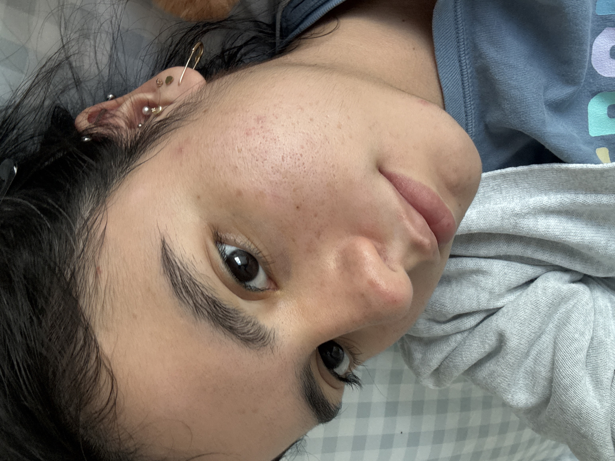 A close-up, high-angle portrait features a person, likely female, with dark hair, lying down and looking directly at the camera. Her head is tilted, giving a unique perspective. Her expression is neutral to subtly smiling.
Visible on her right ear are multiple piercings: a distinctive gold-colored safety pin on the helix, a small flat gold stud, and several silver-toned ball studs and a hoop. Her skin texture is natural, showing visible pores and some minor blemishes on her forehead and cheek. Her eyebrows are dark and appear well-groomed.
She wears a light blue t-shirt, partially visible around her neck and shoulder. Faintly discernible colored letters on the shirt include parts of a purple "R", light blue "E" and "A", and a yellow "D". A soft, grey, possibly knitted or sweatshirt-like fabric is draped around her lower right side. The background shows a light grey and white checkered pattern, suggesting an indoor setting, possibly on a bed. The overall scene conveys a casual, relaxed atmosphere indoors in Austin, United States of America.