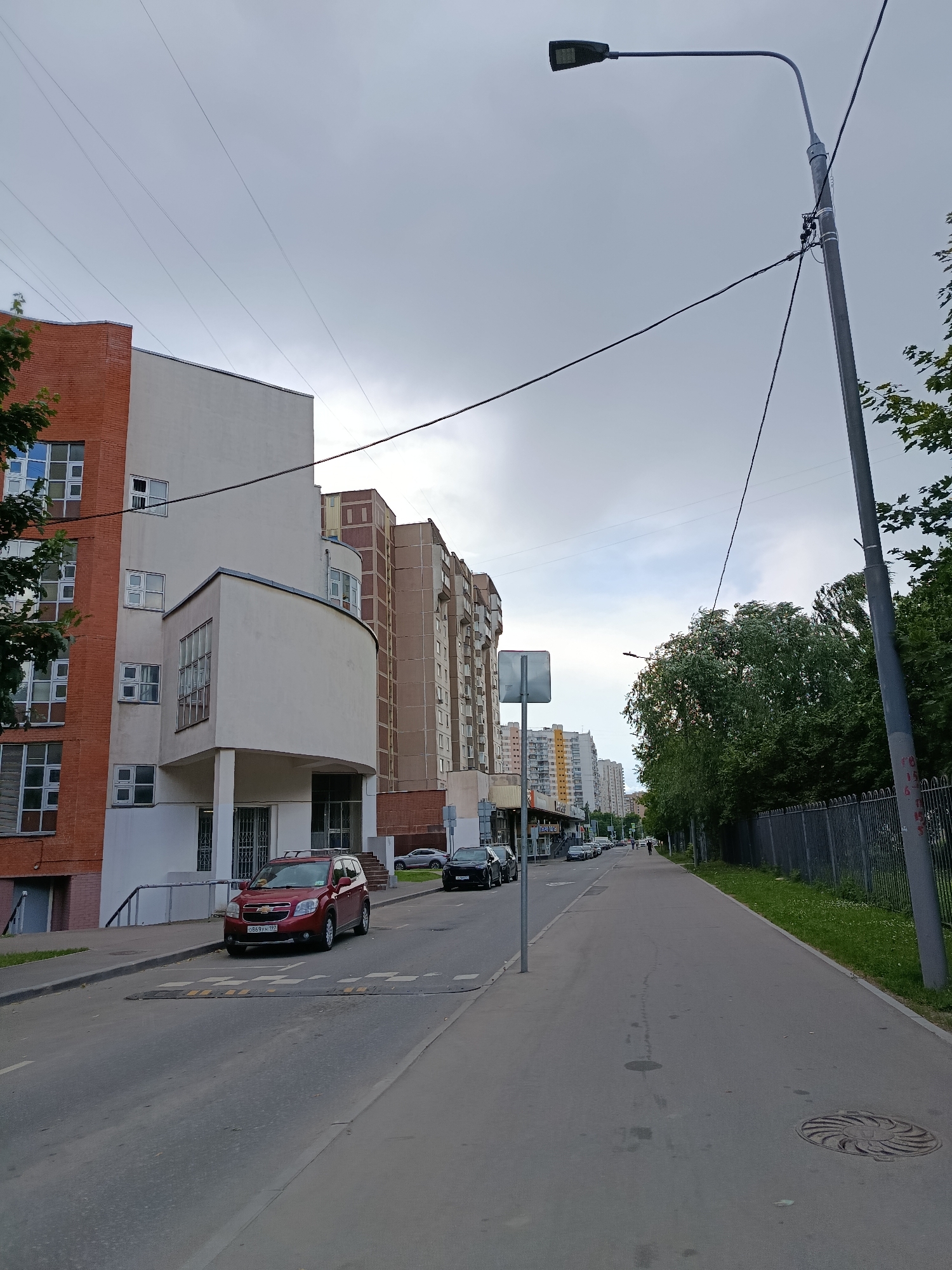 The image depicts a street scene in Moscow, Russia, under an overcast sky. A red Chevrolet Orlando is parked on the street, facing left, behind a speed bump. To the left of the street are multi-story apartment buildings, one with a brick facade and rounded white balconies, and further down, taller buildings with beige and brown exteriors. A sidewalk runs along the right side of the street, bordered by a metal fence and trees. A lamppost stands on the right, casting a shadow on the sidewalk. Several cars are parked or driving on the street in the distance. The scene appears to be a residential area.