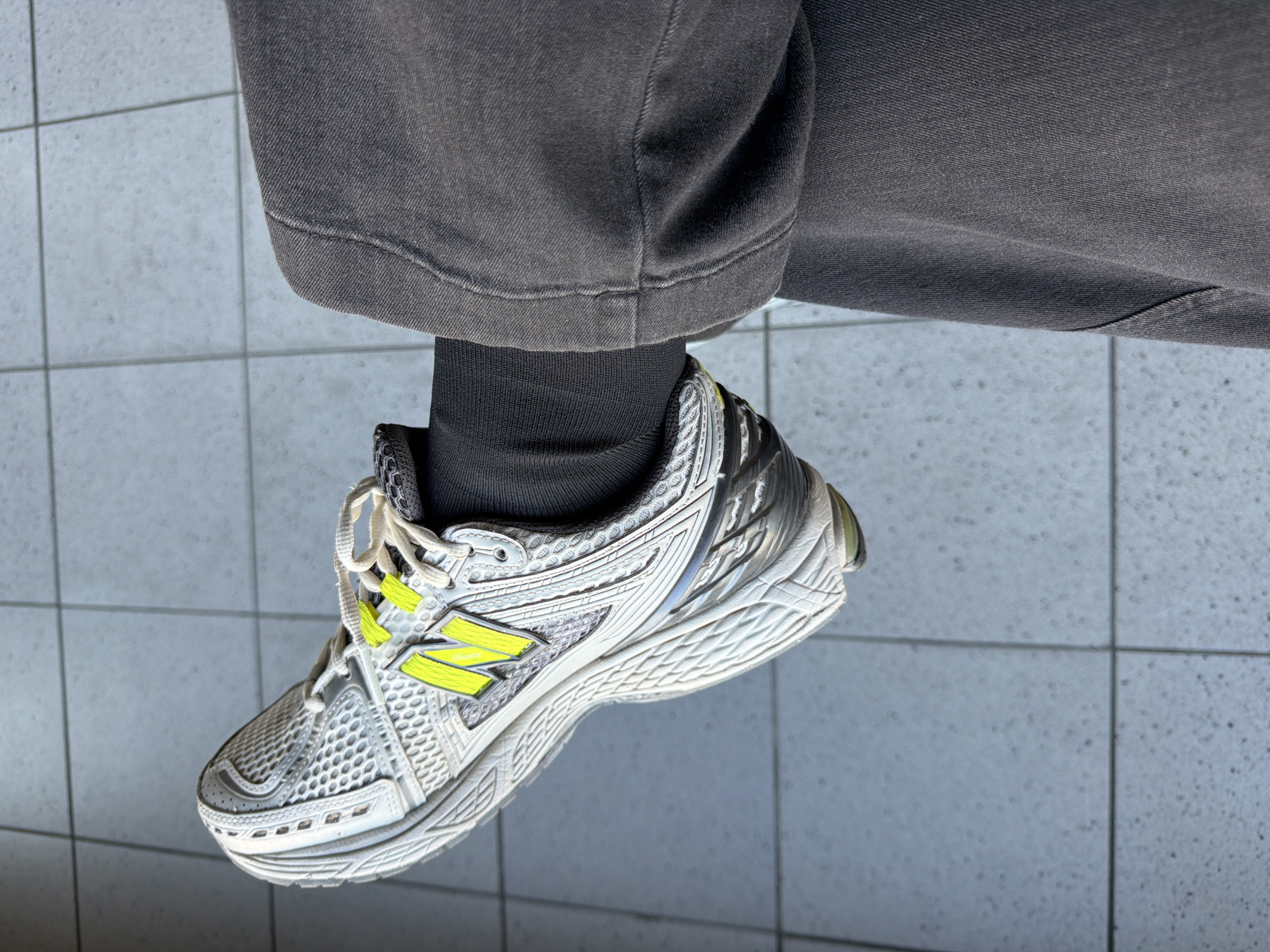 A person's left leg and foot, wearing a grey, washed denim pant and a New Balance 1906R sneaker, is captured in a medium close-up shot. The sneaker is predominantly white with silver accents and bright neon yellow "N" logos on the side. The foot is positioned at a slight upward angle, resting on a tiled floor that appears to be outdoors or in a well-lit indoor space with a blue-grey, speckled tile pattern. The background is blurred, but the distinct tiling suggests a public or semi-public area, potentially a plaza or a walkway in Badhoevedorp, Netherlands, given the context. The lighting is bright, casting subtle shadows and highlighting the textures of the denim and the sneaker's mesh and synthetic materials.