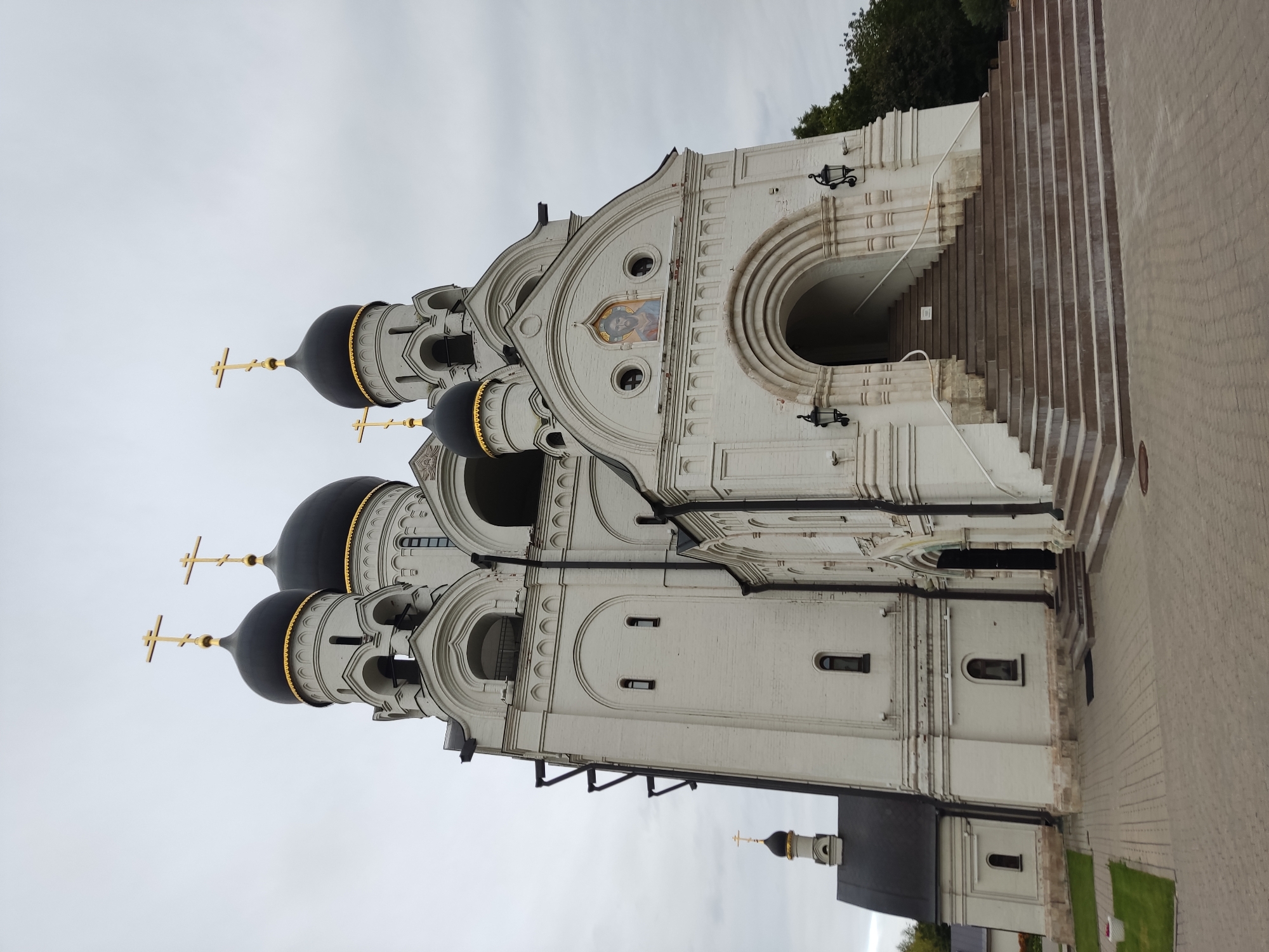 An exterior, low-angle, eye-level, wide shot shows a large, white, multi-domed church under a cloudy sky. The church has five dark, bulbous domes, each topped with a golden cross. A single, smaller dome with a cross is visible on a separate building to the left. The main church structure has arched windows and doorways. A wide set of stairs leads up to the main entrance, flanked by black, ornate lanterns. The paving stones in front of the church are laid in a decorative pattern. Lush green trees are visible to the right of the church.