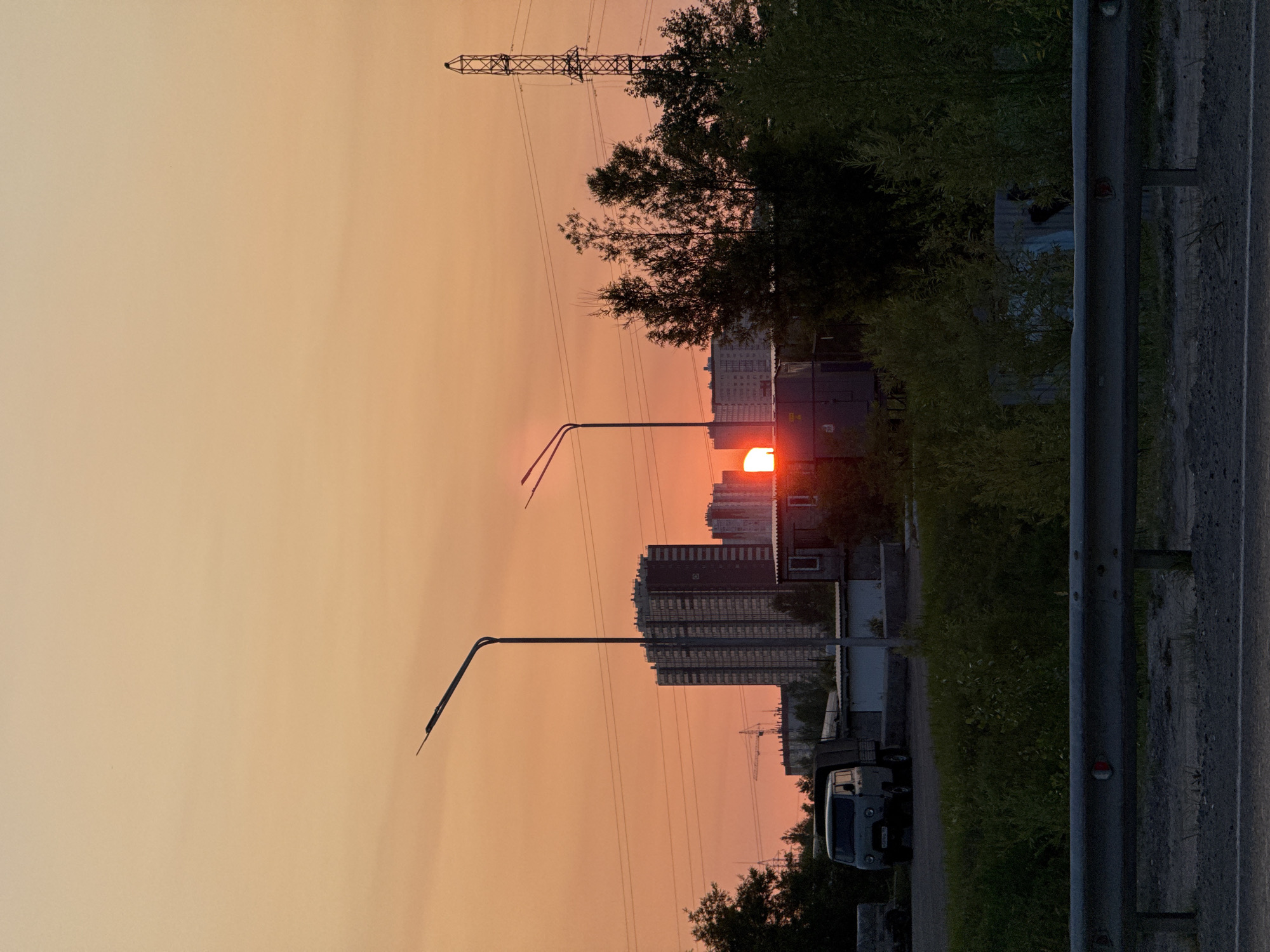 The image captures a sunset in Surgut, Russia. The sky is a gradient of warm orange and yellow hues. In the background, silhouetted against the sky, are several tall, apartment buildings. A large industrial-style building with a crane in front of it is also visible. Two streetlights stand prominently in the foreground, their silhouettes sharp against the sky. A power pylon is located to the right. In the middle ground, there are some smaller buildings and trees. A light-colored van is parked in front of one of the buildings. A metal guardrail runs across the bottom of the image, indicating a road. The overall scene depicts an urban or semi-industrial landscape at dusk.