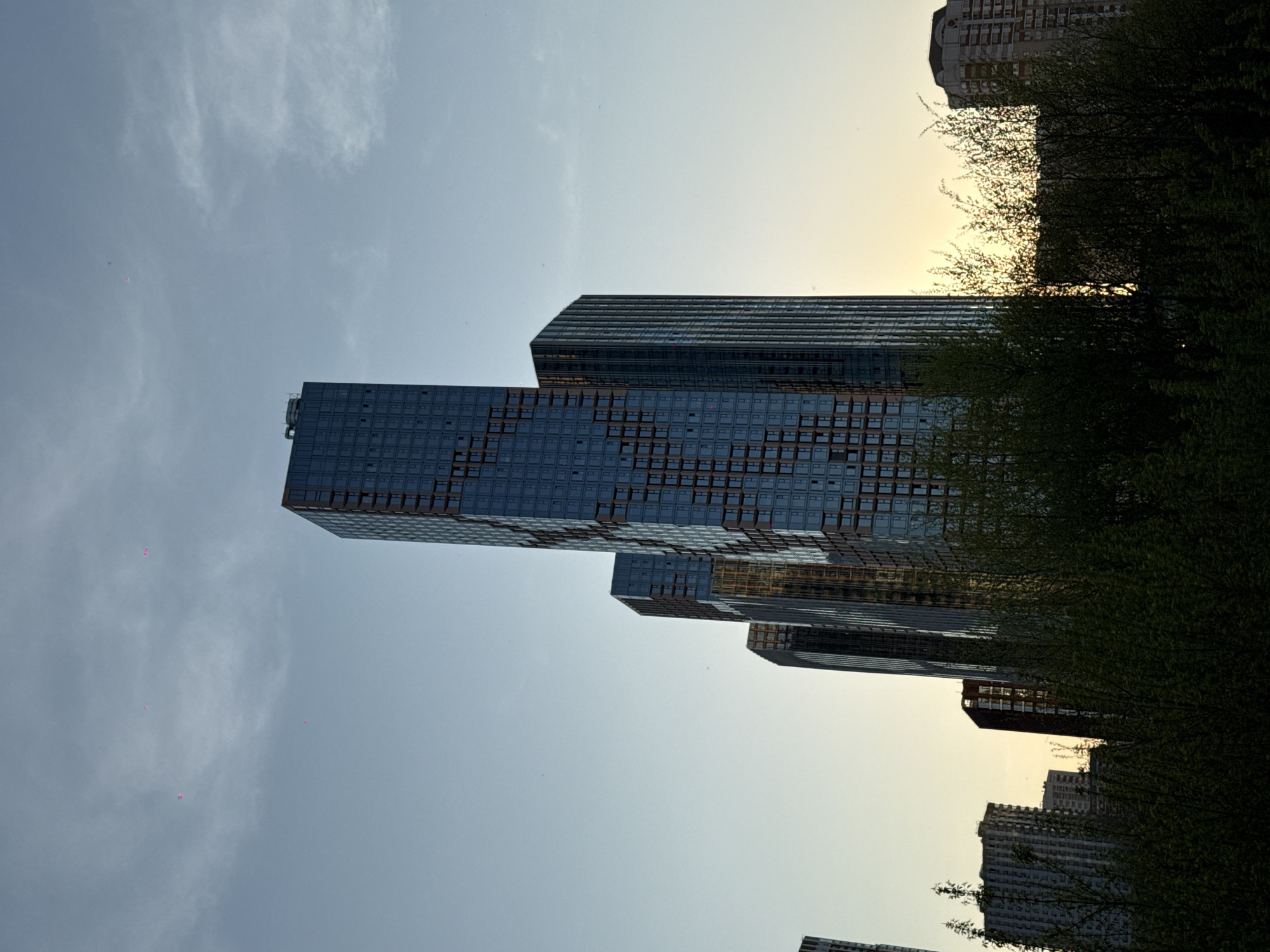 The image is taken from a low angle, looking up at several tall, modern buildings in Moscow, Russia. The buildings have glass facades with a grid pattern of windows and are several stories high. Some buildings are partially obscured by the dense foliage of trees, likely located in a park or green area. The sky is a pale blue with some wispy clouds and a hint of sunset glow visible near the tops of the buildings. There are a few small, pink objects floating in the sky, possibly balloons. The buildings appear to be located in a central area of Moscow.