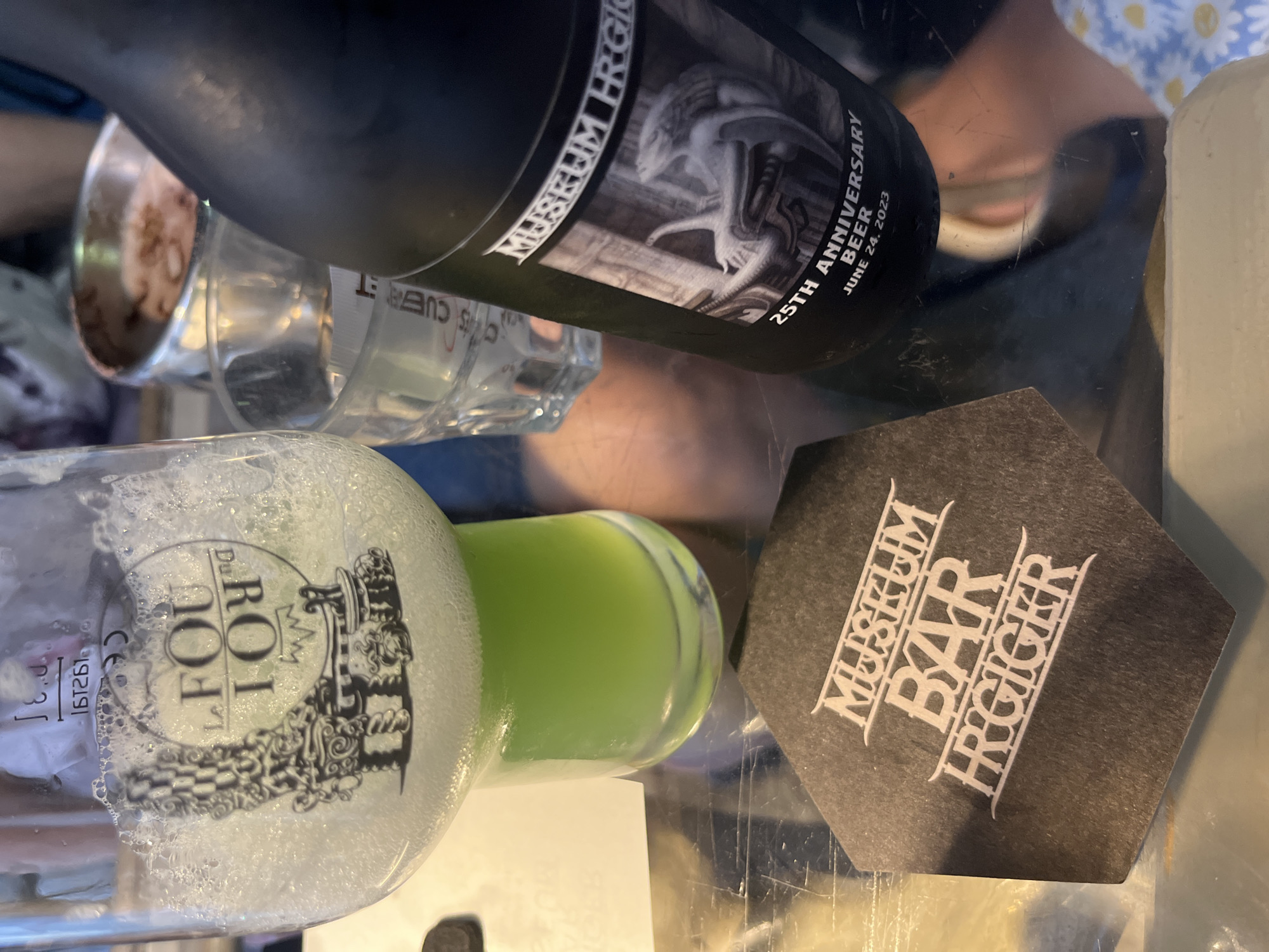 The image shows a table with drinks and coasters. In the foreground, there is a black hexagonal coaster with white text that reads "MUSEUM BAR HRIGER". To the right of the coaster, a black bottle of "MUSEUM HRIGER BEER 25TH ANNIVERSARY JUNE 24, 2023" is standing. The bottle has a label with an illustration of a creature. Behind the bottle, a small glass of clear liquid is visible. To the left, a beer glass filled with a pale green liquid and foam is tilted. The background is blurred and shows a person's arm and a small bowl with a light-colored substance. The image appears to be taken in a bar or restaurant setting. There is no information in the image to suggest a location in Epagny, Switzerland.