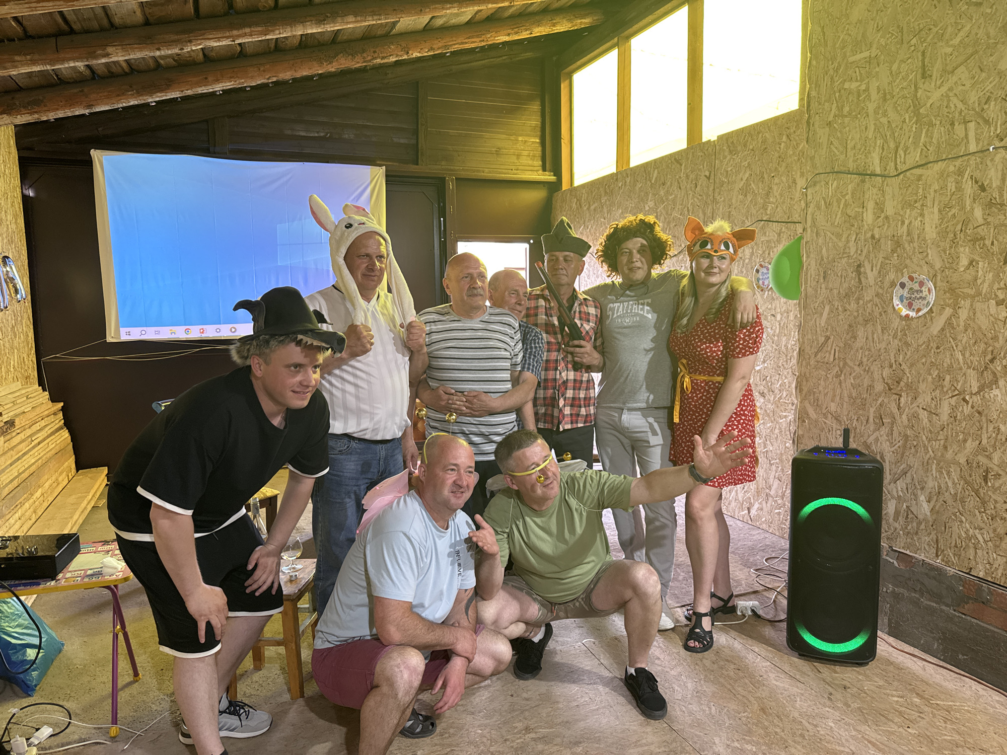 A group of nine people are posing for a photograph indoors. They are dressed in casual clothing, with some wearing humorous costumes or accessories. From left to right, one person is wearing a witch hat and a black outfit, another is wearing a bunny costume, one man is wearing a striped shirt and a yellow party favor around his neck, and two more people are standing behind them, one wearing a red and black plaid shirt and the other one is bald. The sixth person is wearing a grey T-shirt with the word "Stayfree" and an afro wig, and another person is standing to the right wearing a red dress and a fox mask. Two people are squatting in front; one wearing a light blue shirt and a pink party favor around his neck, the other one is wearing a green shirt and a yellow party favor around his nose.
Behind them, there is a projector screen showing a blue image. The walls of the room appear to be constructed of plywood. There is a speaker with green lights on the floor to the right. A small table with items on it is on the left. A portion of a wall with a window can be seen in the top center. The presence of decorations and costumes suggest a party atmosphere. This scene is in the city of Chaikovsky, Russia.