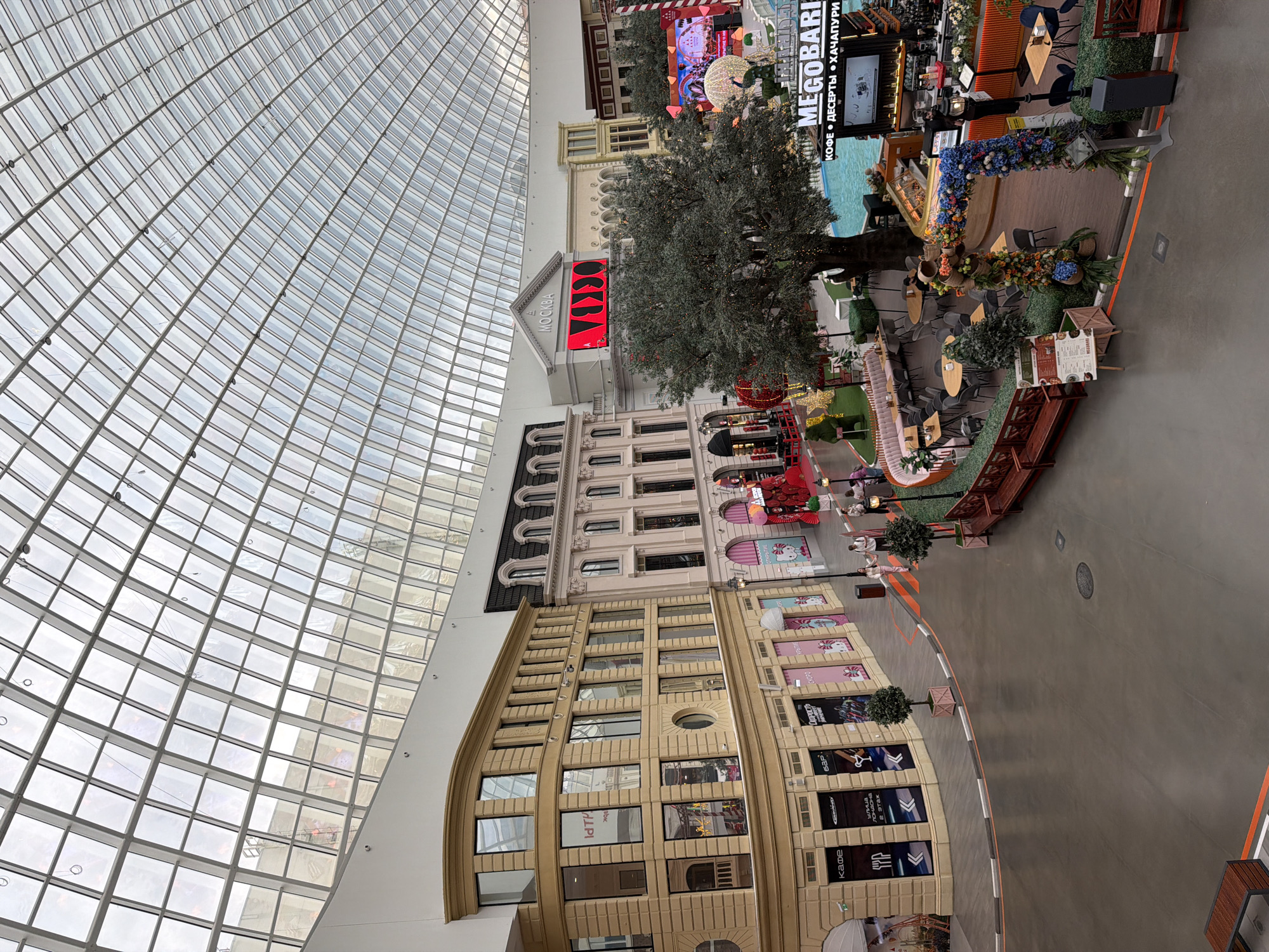The image shows an interior view of a large, modern shopping mall with a distinctive domed glass ceiling. The mall features a European-inspired architectural design with multi-story buildings facades lining a spacious walkway. On the left, a light-colored building with many windows and classical detailing stands. To its right, a taller, darker building with arched windows and a mansard roof is visible. Further in the background, a large screen displaying vibrant graphics is mounted on a building facade that reads "МОСКВА". A large, mature tree with decorative lights is centrally located in front of a body of water, likely a decorative pool or attraction. Several cafe and retail establishments are set up around the pool area, with tables and chairs for patrons. Various decorative elements, including floral arches and themed displays, are scattered throughout the scene. The floor is paved with a smooth, dark material, and orange and white lines mark pathways and borders. A few individuals are present, walking and sitting at tables, suggesting moderate foot traffic. The overall impression is one of a vibrant and engaging indoor entertainment and shopping complex.