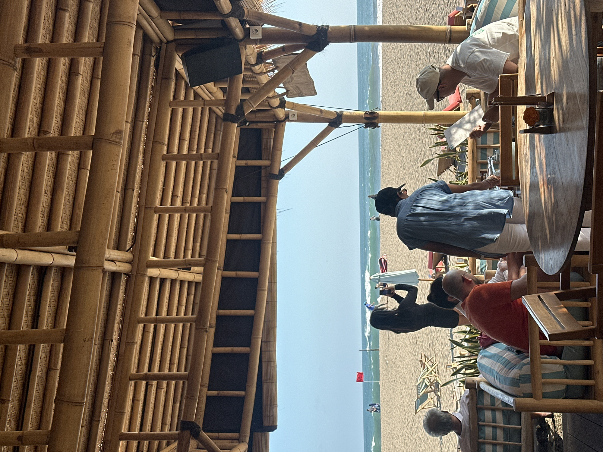The image captures a scene at a beachside restaurant in North Kuta, Indonesia. The foreground features a round wooden table with a small vase containing orange flowers and a menu. Several people are seated or standing in the mid-ground, beneath a roof constructed of bamboo poles and woven bamboo mats. One person in a white t-shirt and baseball cap is seated and reviewing a menu. Another person wearing a blue shirt and a dark baseball cap is standing and appears to be interacting with the seated person. Behind them, another individual in an orange shirt is seated. A person in a black outfit is standing further back, facing the ocean and holding up a phone, possibly taking a picture. The background shows a sandy beach with lounge chairs and a red flag planted in the sand. The ocean with moderate waves is visible beyond the beach, meeting a clear blue sky. The overall ambiance suggests a relaxed, casual dining experience by the sea.