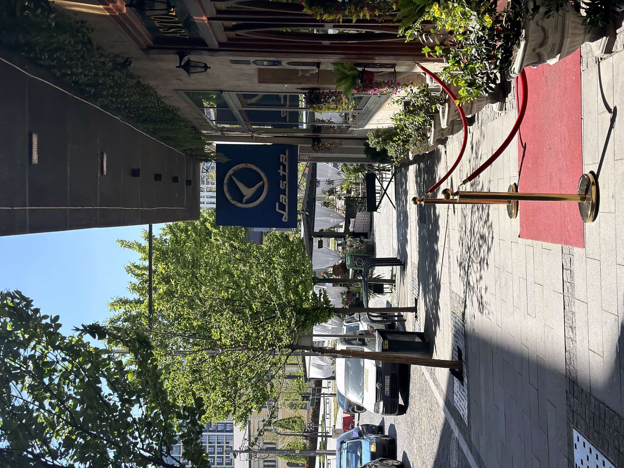 The image shows a street scene in Belgrade, Serbia, with a restaurant entrance and outdoor seating area. A red carpet and gold stanchions are visible, marking the entrance to the "ASTORIA" restaurant. A blue sign with a white bird logo reads "ASTORIA." Outdoor seating with tables and chairs is visible under white canopies. Cars are parked along the street, and trees line the sidewalk. Buildings can be seen in the background. The weather appears sunny and clear. The image has been rotated 90 degrees counterclockwise.