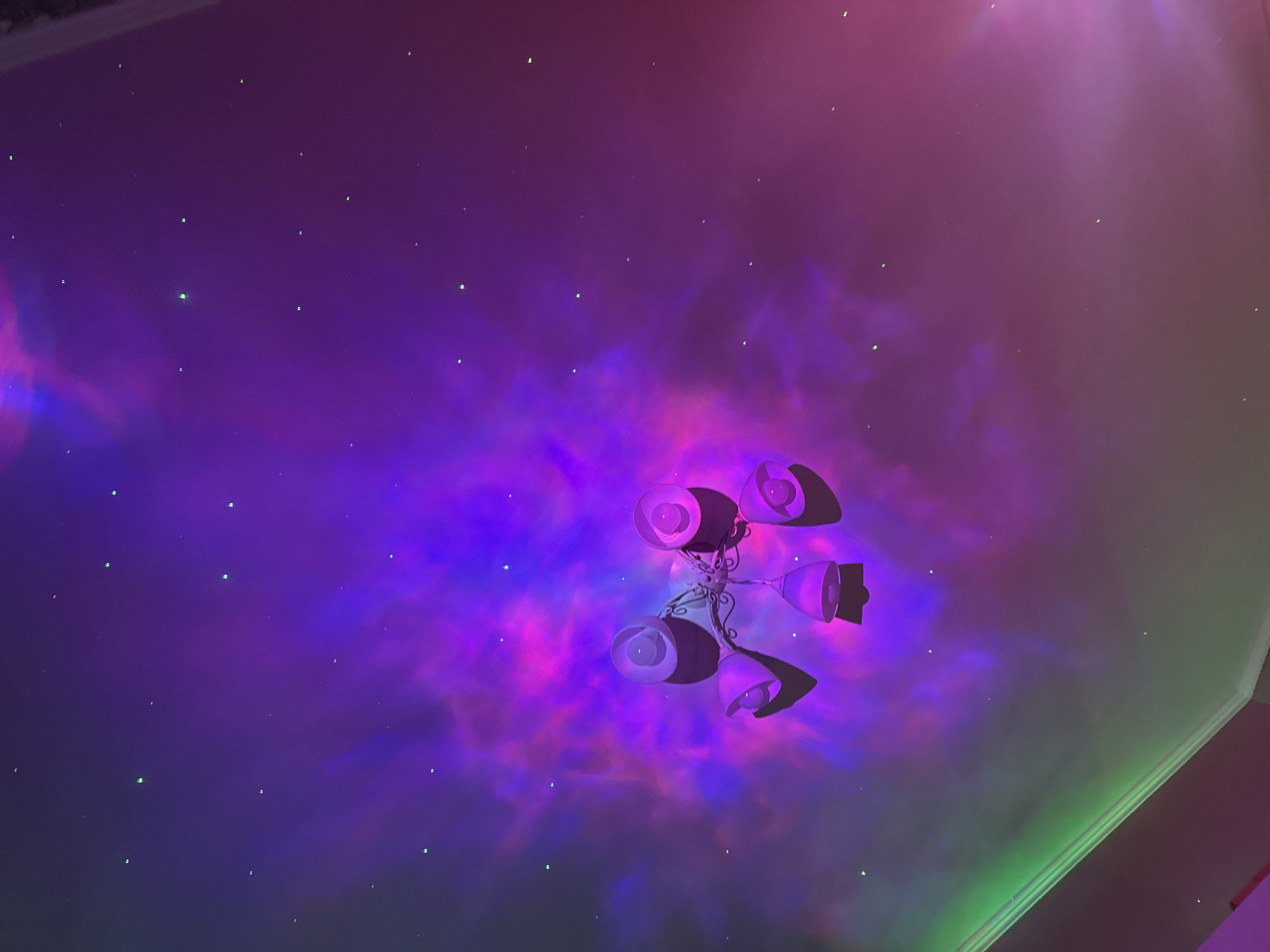 The image is a low-angle shot of a ceiling in an indoor environment. The ceiling is bathed in purple, blue, and red light, simulating a nebula or galaxy effect, with small green dots scattered across it, resembling stars. A white, ornate ceiling light fixture with multiple lamps is mounted at the center of the image. The light fixture is casting shadows on the ceiling. The right side of the image shows a section of a wall or architectural detail in green and pinkish-red colors. The location and city are unknown.