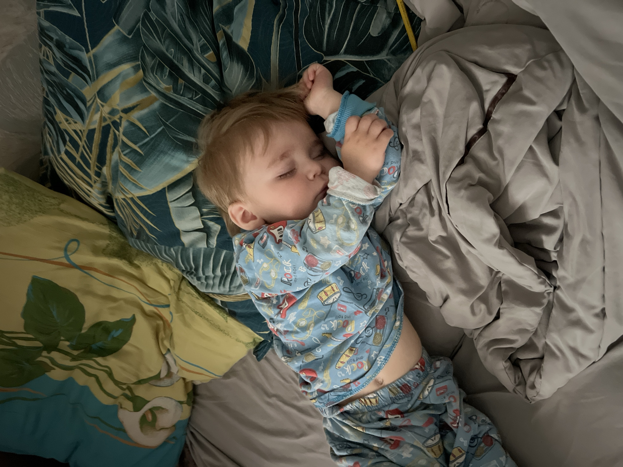 A young child is sleeping on a bed. The child is wearing blue pajamas with a "Rock and Roll" pattern. The child is lying on their back with their hands near their face. The bedding includes pillows with floral and leaf patterns, and a gray blanket. The location is not specified in the image.