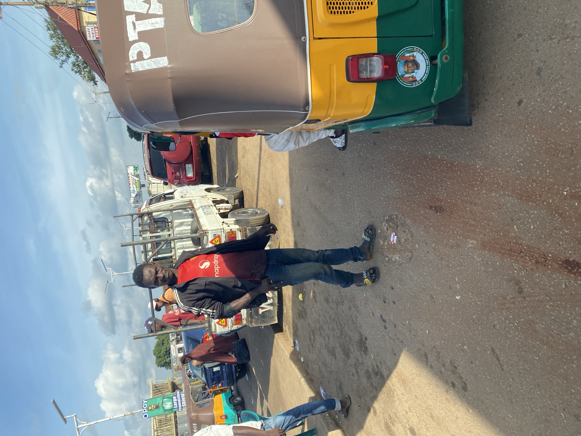 A man wearing a red t-shirt with a white logo and a black jacket over it, dark blue jeans, and black sandals with a yellow stripe stands on a dirt road in Gura Top, Nigeria. He is looking at the camera with a neutral expression. Behind him, several vehicles are visible, including a white truck, a red SUV, and a yellow and green auto-rickshaw. In the background, there are buildings and a blue sky with scattered clouds. The overall scene appears to be a street market or a busy roadside. The auto-rickshaw has a sticker on its rear with a portrait of a man and text that reads "OMAR MUSA YAR'ADUA" and "FEDERAL REPUBLIC OF NIGERIA".