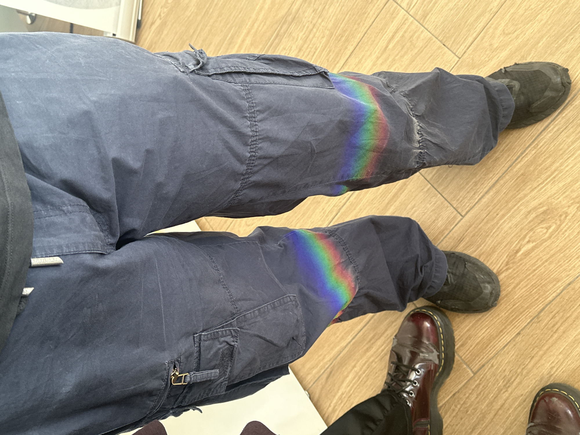 The photograph shows two pairs of legs clad in cargo pants and shoes, positioned on a light brown, wood-like floor. The top pair of legs wears navy blue cargo pants and dark gray shoes. A rainbow-like refraction of light is visible on the left leg of the top pair of pants. The bottom pair of legs wears navy blue cargo pants and burgundy, patent leather Doc Martens. The location appears to be indoors. There is no indication this image was taken in Quart de Poblet, Spain.