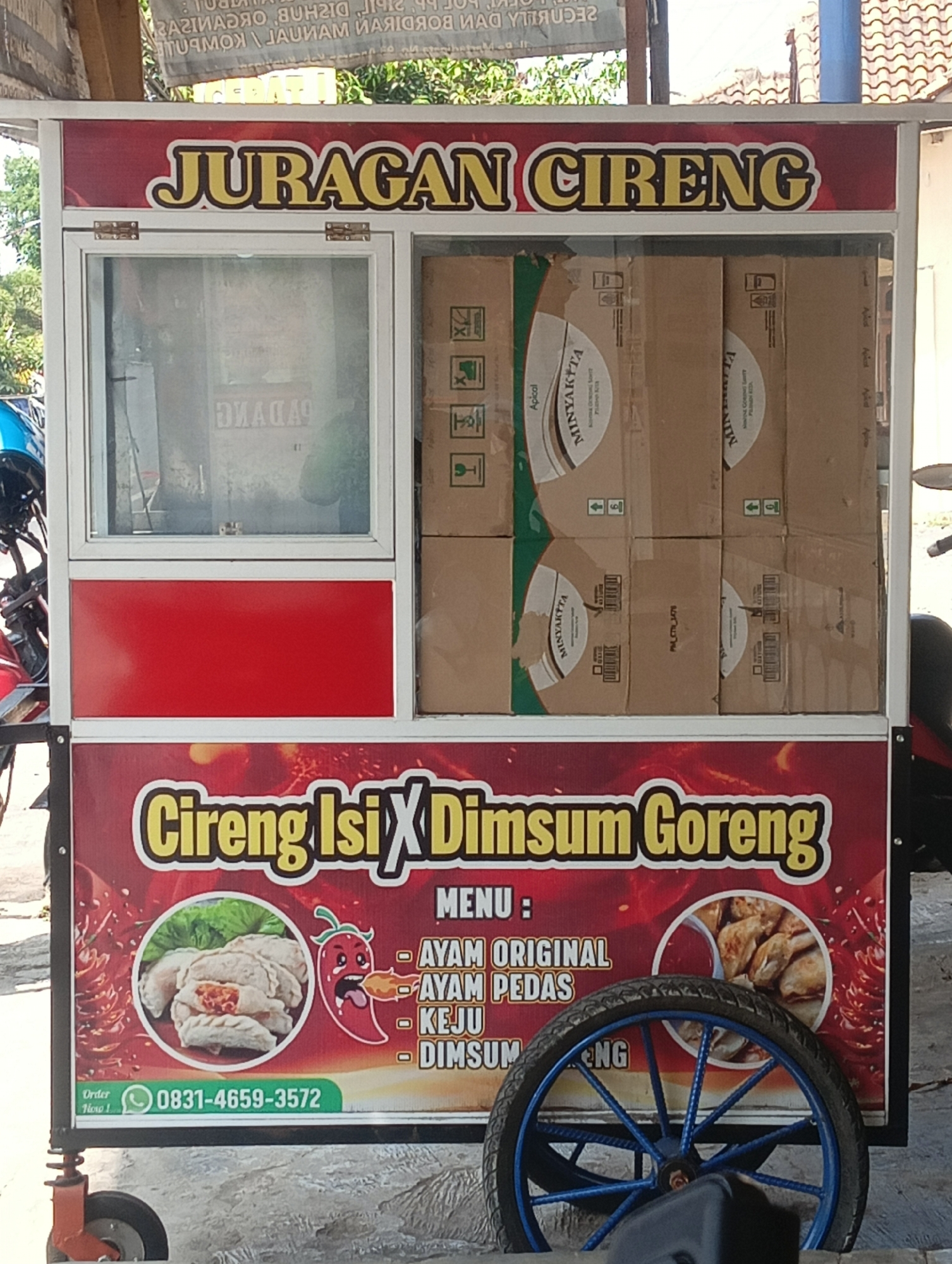 The image shows a food cart selling "Cireng Isi Dimsum Goreng" in Manggari, Indonesia. The cart is red and white with a large banner at the bottom displaying the name of the food and a menu. The menu lists "Ayam Original," "Ayam Pedas," "Keju," and "Dimsum Goreng." There are also pictures of the food on the banner, as well as a chili pepper graphic. The cart has a glass display case on top, filled with boxes of product. A bicycle wheel with blue spokes is attached to the front of the cart. The cart is parked on a street, with buildings and foliage visible in the background.