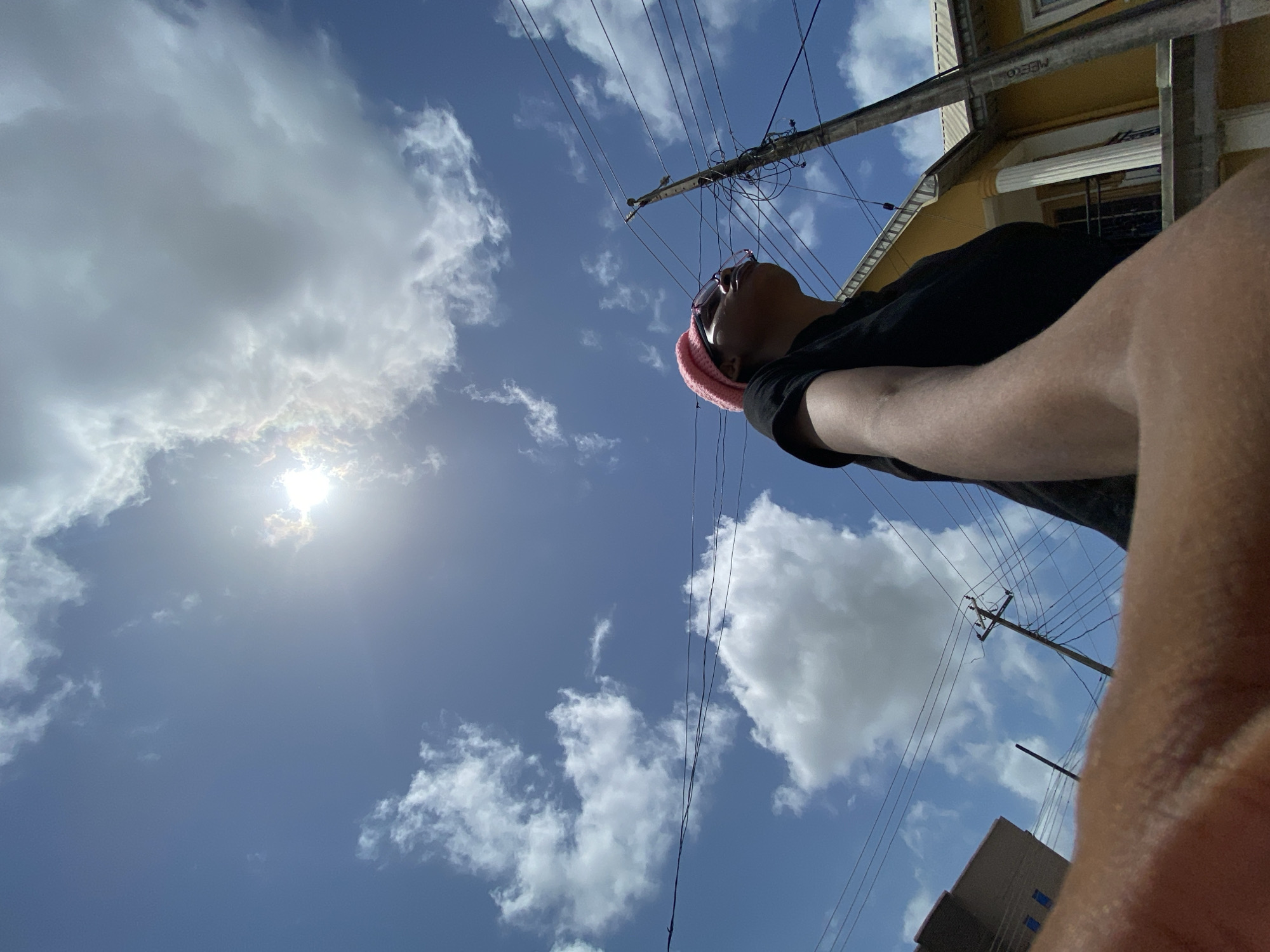 The image shows a person looking up at a partly cloudy sky. The sun is visible through the clouds. Utility poles and wires are present. A building is visible in the background. The location is in a city of Unknown, Unknown.