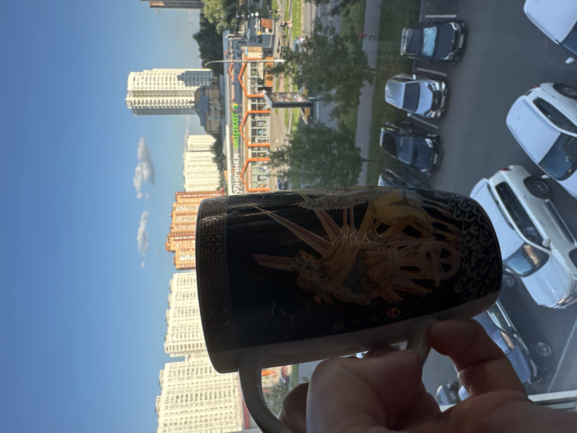 The image shows a close-up of a hand holding a black mug with a decorative pattern, with a city skyline in the background. The mug features a depiction of a chariot with a figure driving it, along with what appears to be a sunburst or radiating lines. The background consists of several multi-story residential buildings, some appearing to be of white and orange brick construction. A supermarket with the word "MARKET" prominently displayed in green letters is visible, along with other commercial establishments. A parking lot with several cars is also present. The sky is blue with a few scattered clouds. The overall setting suggests an urban environment. The city of Podolsk, Russia, is identified as the location.