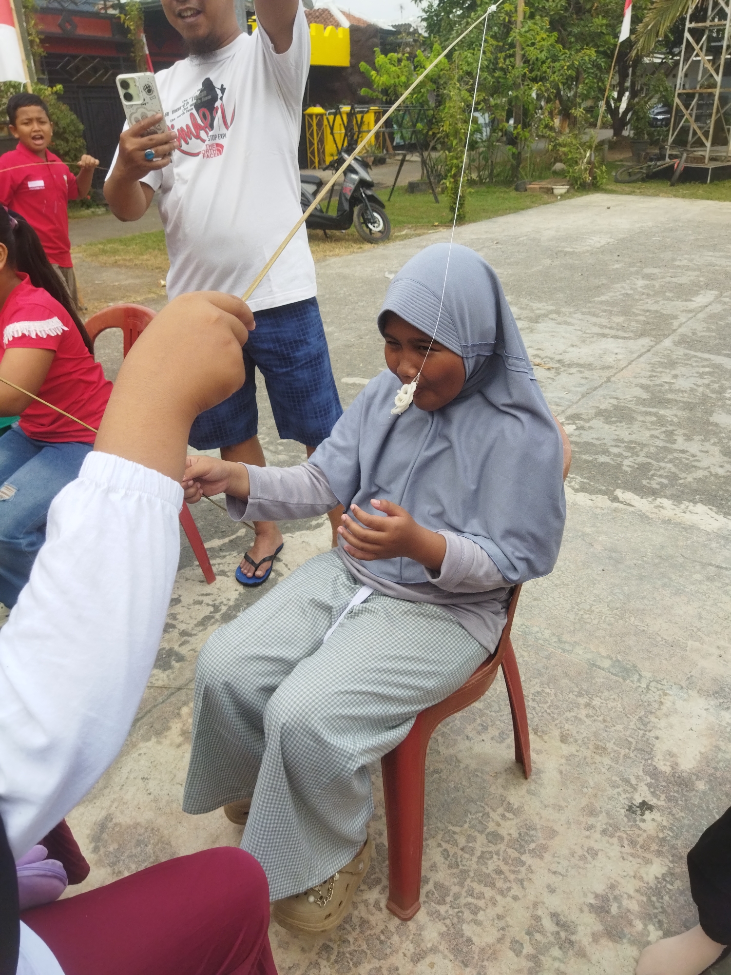 A young Indonesian girl wearing a grey hijab and grey and white checkered pants sits on a red plastic chair outdoors, participating in a game. A thin white string is attached to a white puff snack, which is dangling in front of her mouth. She is attempting to eat the snack without using her hands, which are held down. Her right hand is extended towards the snack, and her left hand is resting in her lap.
Behind her, an adult male wearing a white t-shirt and blue checkered shorts is holding a smartphone in his left hand. A younger boy in a red shirt is standing behind him, also holding a stick. To the left, partially visible, is the arm of another person wearing a white long-sleeved shirt. The ground is made of concrete with a speckled pattern. In the background, there are trees, a motorcycle, and a building with a yellow facade and red trim. Indonesian flags are visible on poles. The scene appears to be a community event or celebration, possibly in the city of Banjar, Indonesia, given the presence of Indonesian flags and cultural attire.