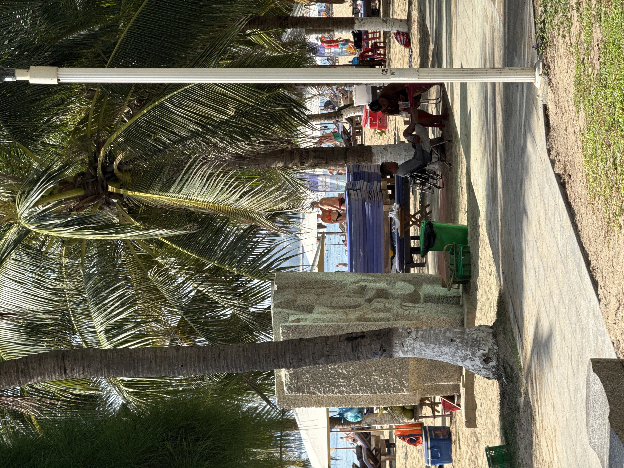 The image depicts a sunny day on a beach in Nha Trang, Vietnam, likely near a resort or public area. In the foreground, a paved walkway runs along the edge of the sandy beach. A large, light-colored stone sculpture, possibly abstract art, stands prominently near the center of the frame, with a textured surface and a palm tree trunk obscuring part of it. Several palm trees with lush green fronds cast shadows on the sand.
Various beach amenities are scattered throughout the scene. Beach chairs and loungers are visible, some occupied by people. Blue plastic containers, a red cooler, and green baskets are placed on the sand, suggesting a relaxed beachgoer atmosphere. In the middle ground, a row of stacked blue cushions or mats is set up, possibly for seating or a lounge area. Further back, people are seen near the water, some in swimwear, and beach umbrellas offer shade.
In the immediate foreground, two men are seated at a low table under the shade of a palm tree. One man, wearing a grey t-shirt and shorts, is looking at his phone, while the other, shirtless and wearing red shorts, is looking down at the table. A white lamppost stands to the right of the frame, a common feature in public beach areas. The ocean is visible in the distance, with calm water and a few boats or structures on the horizon. The overall impression is a vibrant and active beach scene under a clear sky.