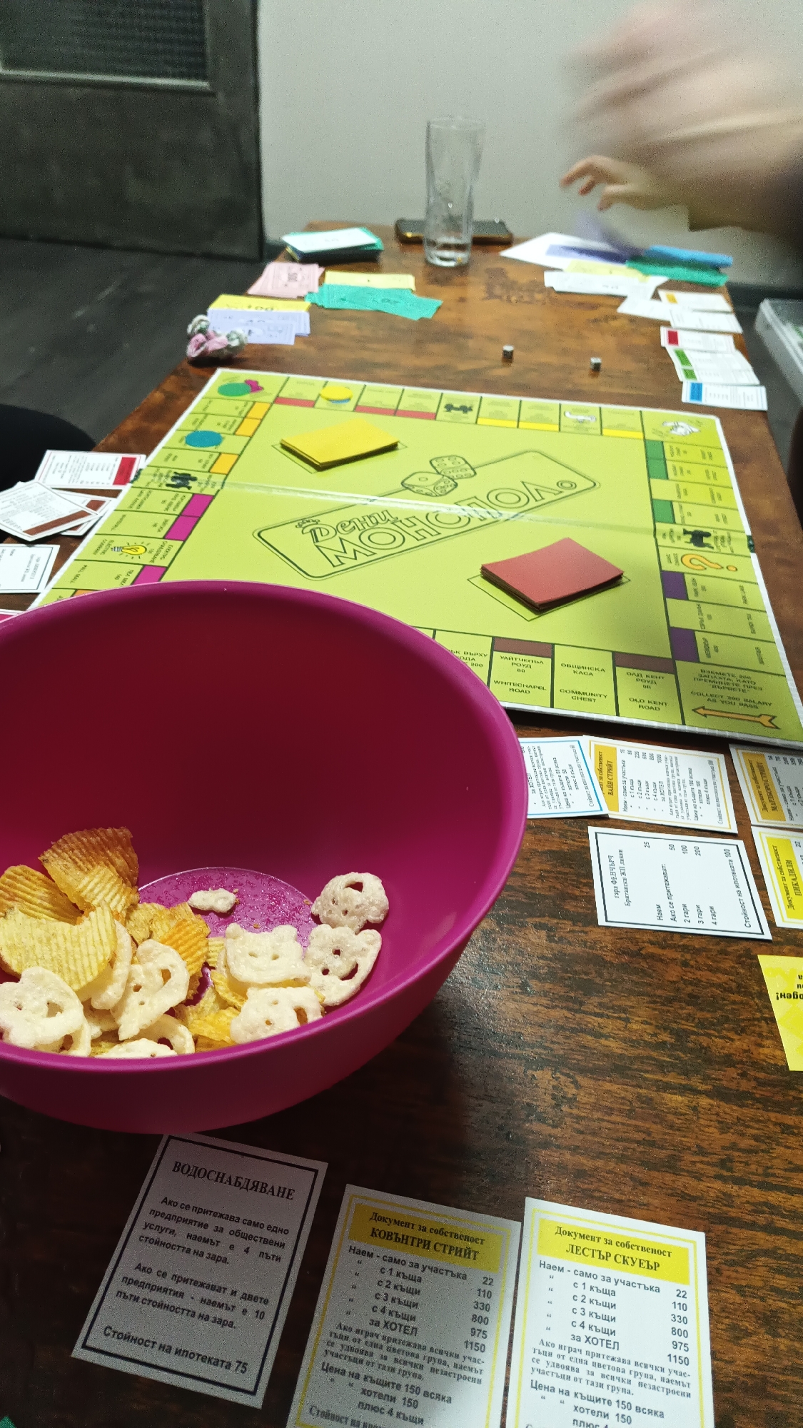 A top-down view captures a lively game of Monopoly being played on a wooden table. The central focus is the bright green Monopoly board, which features Bulgarian text, prominently displaying "МОНОПОЛ" (Monopol) in the middle. Stacks of colorful Monopoly money and various property cards are spread across the table. Two small metallic dice rest on the board, and a yellow stack of "Chance" or "Community Chest" cards is visible.
To the left, a large pink bowl contains an assortment of snacks, including ridged potato chips and white, animal-shaped crackers, indicating a casual setting. Several property cards are visible in detail, including "ВОДОСНАБДЯВАНЕ" (Water Supply), "КОВЪНТРИ СТРИЙТ" (Coventry Street), and "ЛЕСТЪР СКУЕЪР" (Leicester Square), all with Bulgarian text detailing rent values and house/hotel costs. Other partially visible cards include "ВАЙН СТРИЙТ" (Vine Street) and "ПИКАДИЛИ" (Piccadilly), along with railway property cards like "ГАРА ФЕНЧЪРЧ СТРИЙТ" (Fenchurch Street Station).
In the background, a blurry human hand reaches across the table towards some cards, suggesting active gameplay. A clear glass, partially filled with liquid, and a dark-colored smartphone are also on the table. The setting appears to be indoors, likely an evening gathering, with a dark door or wall visible in the background. The scene depicts a typical board game night with snacks and friends.