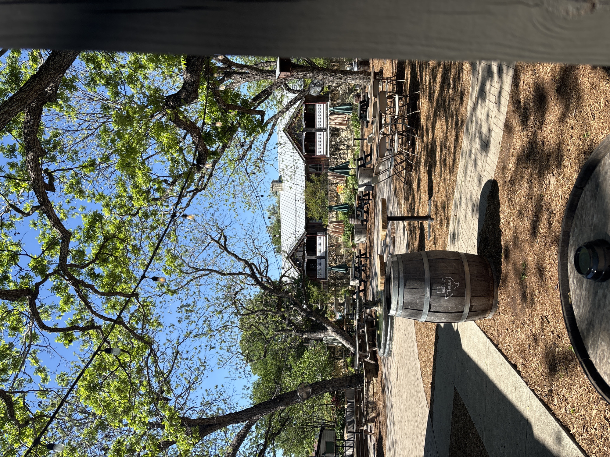 The image shows an outdoor patio area in New Braunfels, Texas. Two two-story buildings with white metal roofs and brown siding are visible beyond the patio. A brick walkway leads through the patio area, and wooden tables and chairs are set up for outdoor dining. A wooden barrel with the words "Dry Comal Creek Vineyards" is in the foreground. Trees with green leaves and string lights overhead provide shade. The sky is blue.