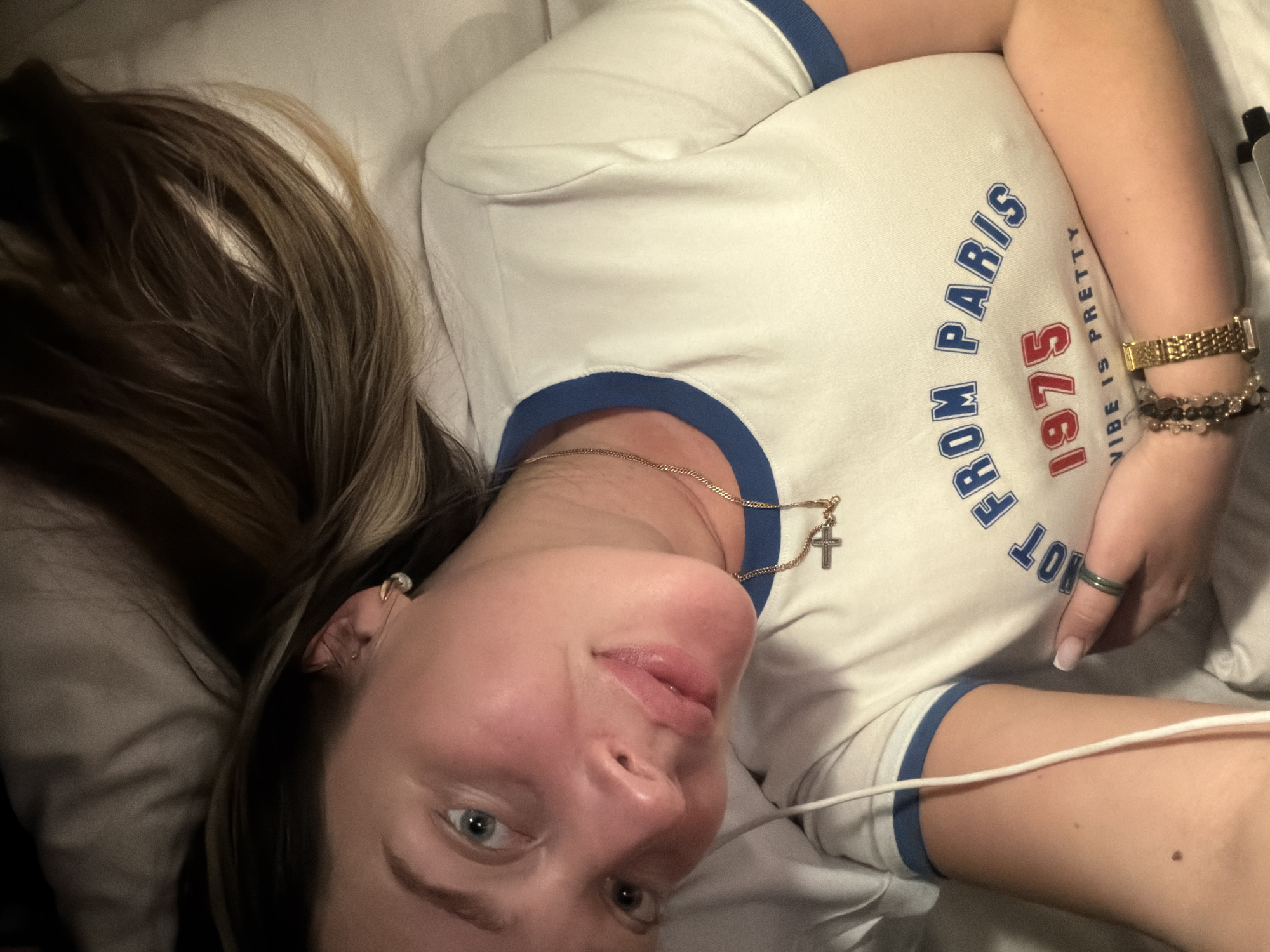 A woman is lying on a white pillow in what appears to be a bed. She is wearing a white T-shirt with blue trim and the words "FROM PARIS 1975 VIBE IS PRETTY" printed on it. She has brown and blonde hair, and is wearing a gold necklace with a cross pendant. She is also wearing a gold watch, and a ring on her finger. A white wire is visible near her arm. The location is not discernible from the image. There is no indication that this image was taken in Moscow, Russia.