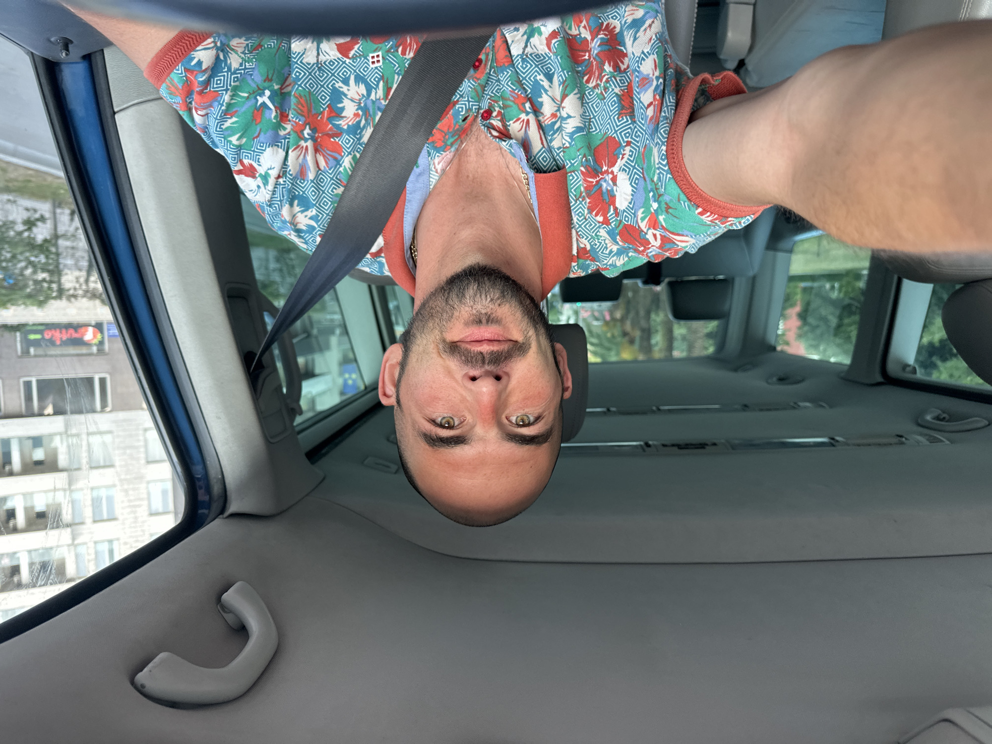 A man is driving a vehicle in Budva, Montenegro. He is a bald man with a beard and is wearing a colorful shirt. The man is looking at the camera and is wearing a seatbelt. The interior of the vehicle is visible, including the dashboard, steering wheel, and seats. The exterior of the vehicle shows buildings and trees.