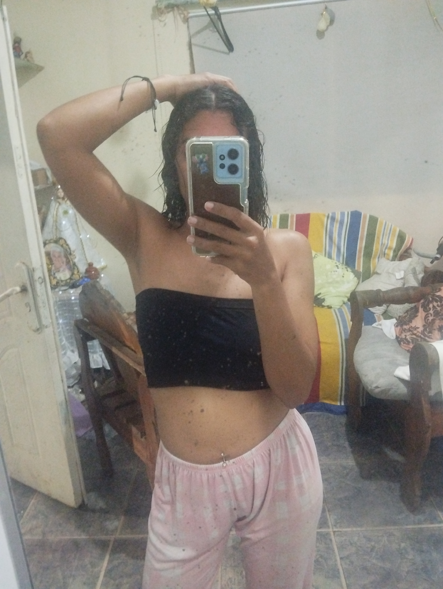 A woman stands in front of a mirror taking a selfie with a smartphone. She wears a black tube top and pink pajamas. Her hair appears wet. Behind her, there is furniture including a striped couch and a wooden chair. The location is likely El Valle del Espíritu Santo, Venezuela, based on the prompt instructions. The tile floor and door are visible in the foreground.