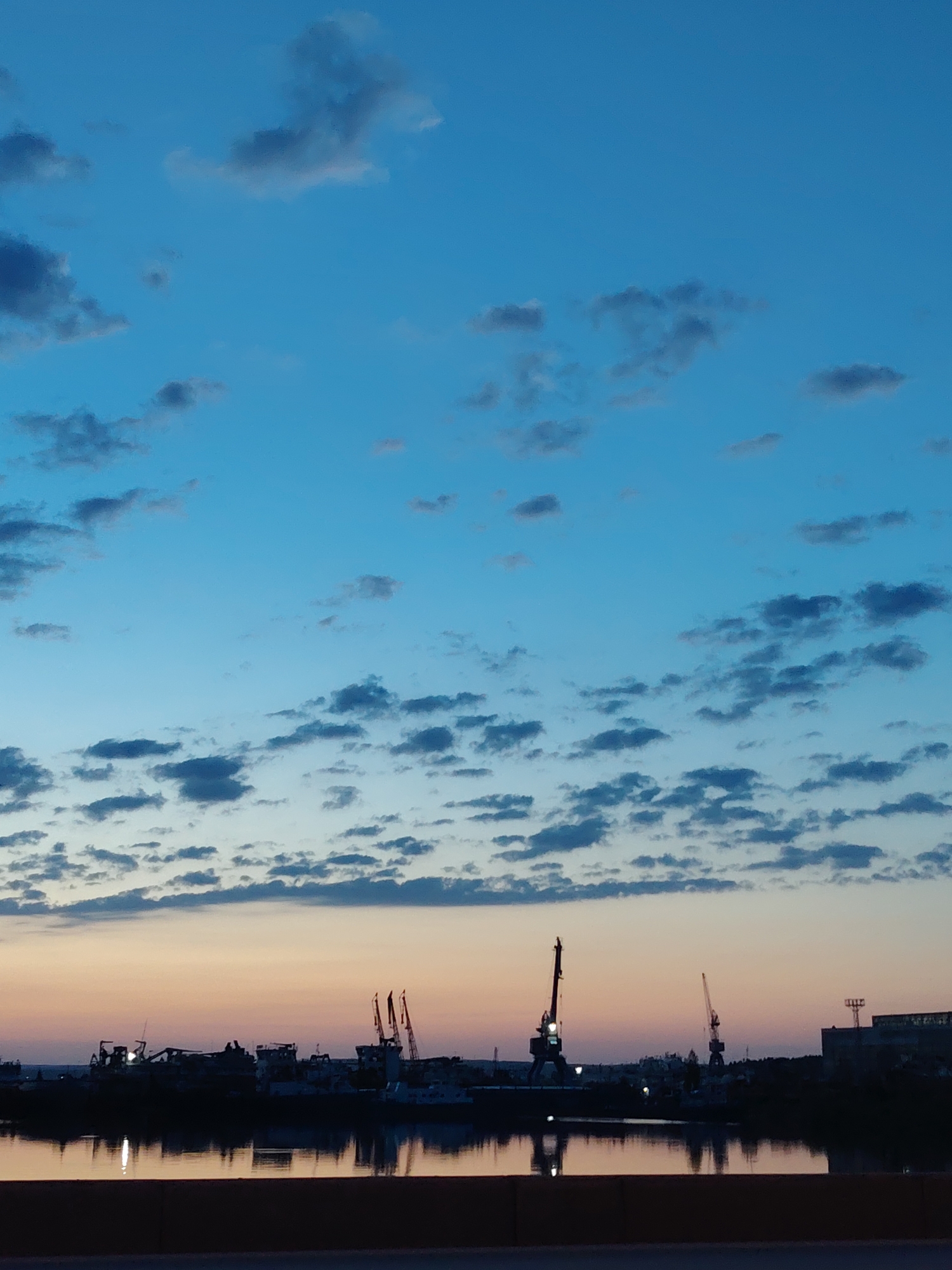 The image shows a waterfront scene in Chaikovsky, Russia, at dusk or dawn. The sky transitions from a darker blue at the top to a lighter orange/yellow near the horizon, with scattered clouds throughout. Below the horizon line, there is a body of water reflecting the sky and the lights of the structures along the shore. Silhouetted against the colorful sky are several cranes and buildings, indicative of a port or industrial area. A dark foreground element, possibly a bridge or a barrier, is at the very bottom of the frame. No people are visible in the image.