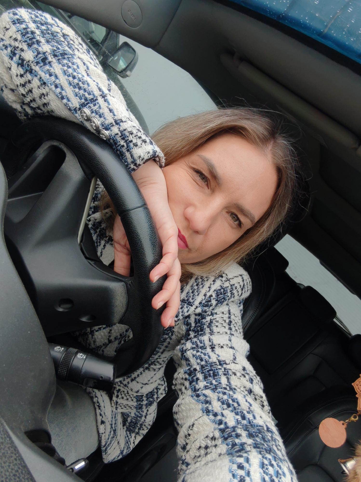 A woman sits in the driver's seat of a car, resting her head on her hand, which is placed on the steering wheel. She is wearing a blue and white patterned jacket. The interior of the car is visible, including the steering wheel, dashboard, and seats. The background appears to be an overcast sky with some reflections on the windshield, suggesting it may be raining or the sky is cloudy. No specific landmarks of Petropavlovsk-Kamchatsky, Russia are identifiable in this image.