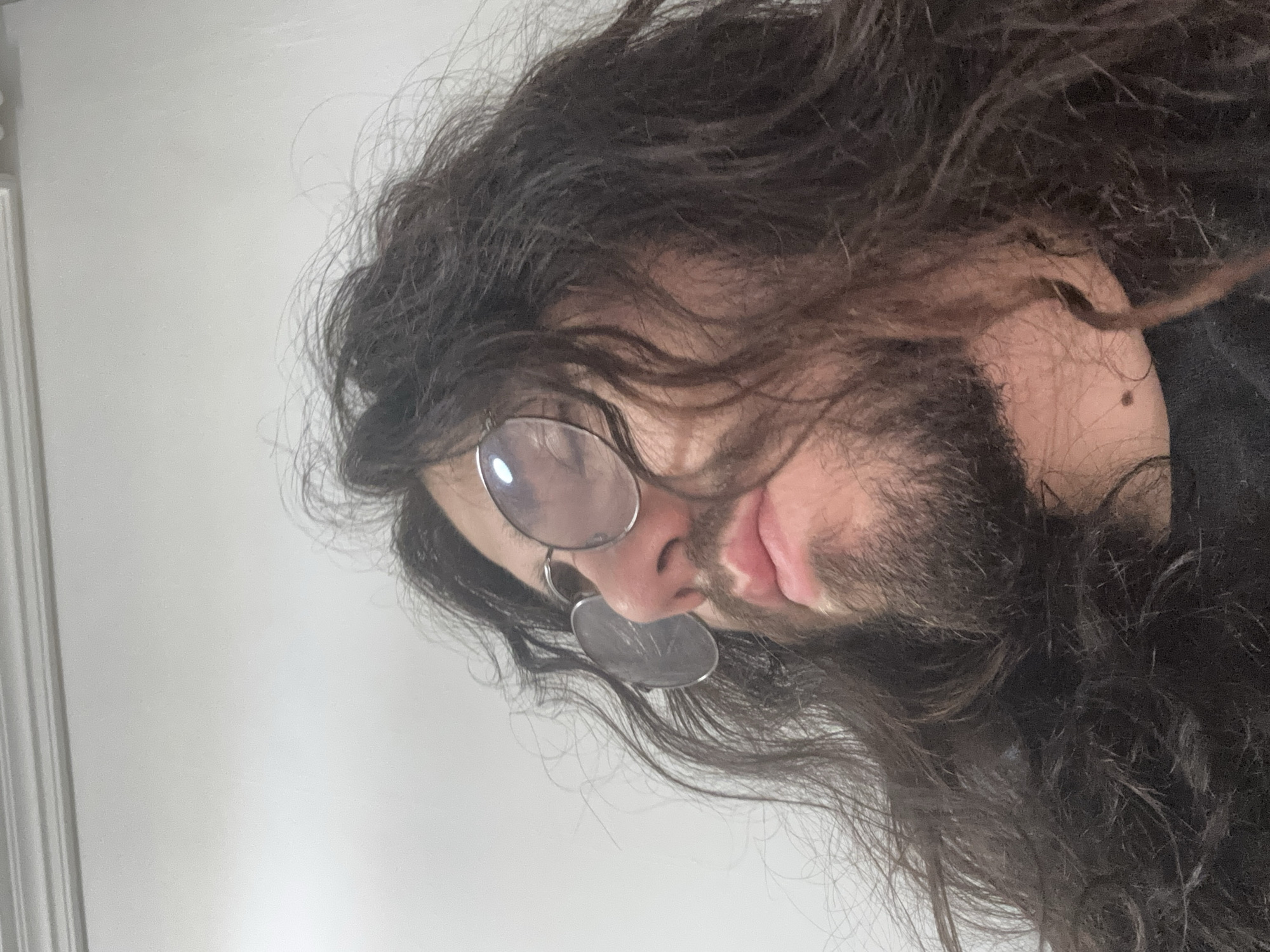 An extreme close-up shot shows a person's face from a low angle, with the head tilted upwards and to the left. The person has long, dark, wavy hair that partially obscures their face. They are wearing round, thin-rimmed glasses. The right lens of the glasses reflects a bright light source, while the left lens is less reflective. The person's nose and mouth are visible, and they have a beard. The background is a plain, light-colored wall with a decorative molding at the top. The scene appears to be indoors, possibly in an office or a room. No city or specific location information is available.